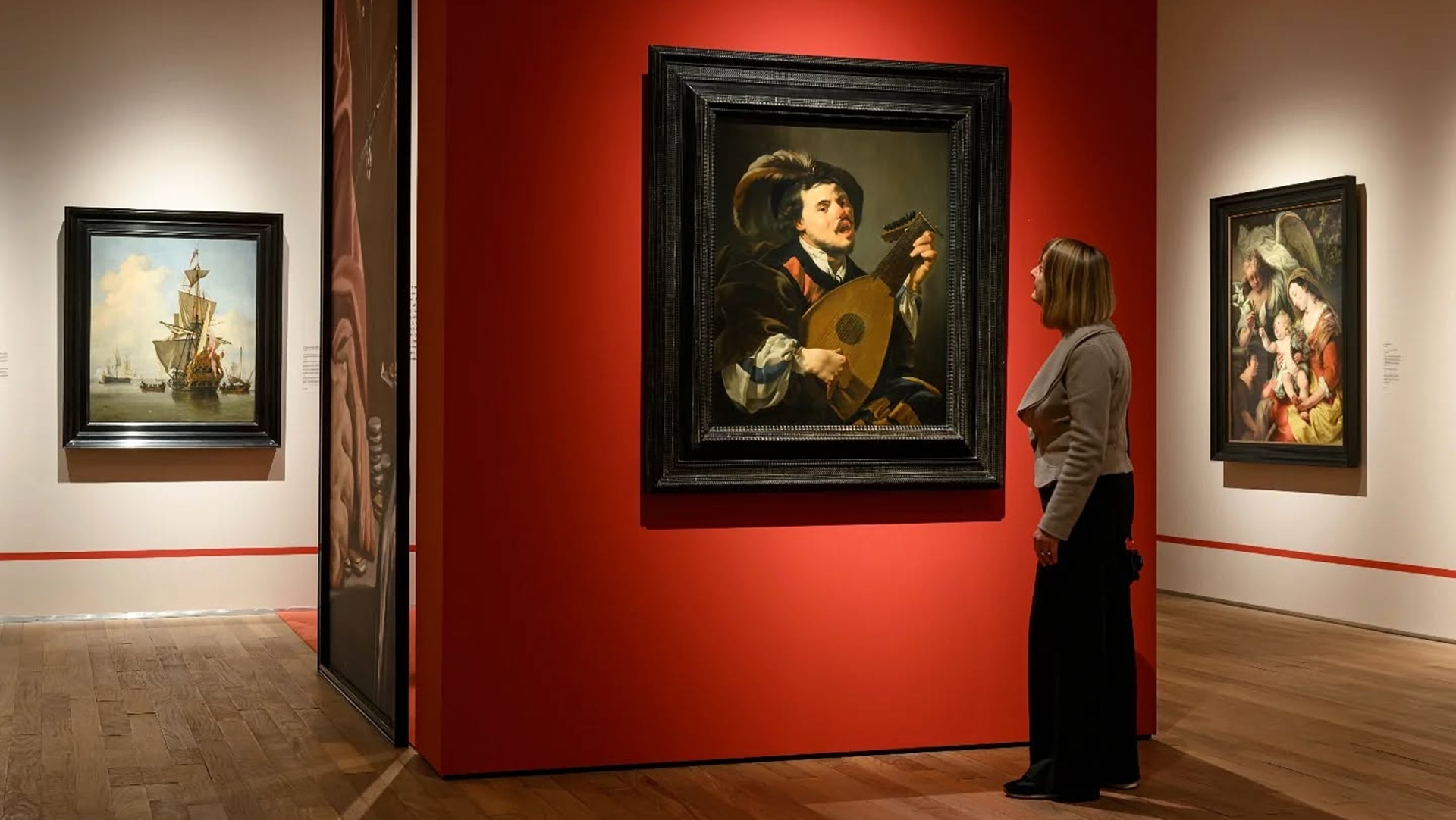 Een vrouw kijkt naar een schilderij van een muzikant met een luit in een museum, met een rode muur als achtergrond. Andere schilderijen, waaronder een afbeelding van een schip en een religieuze scène, zijn ook te zien in de ruimte.