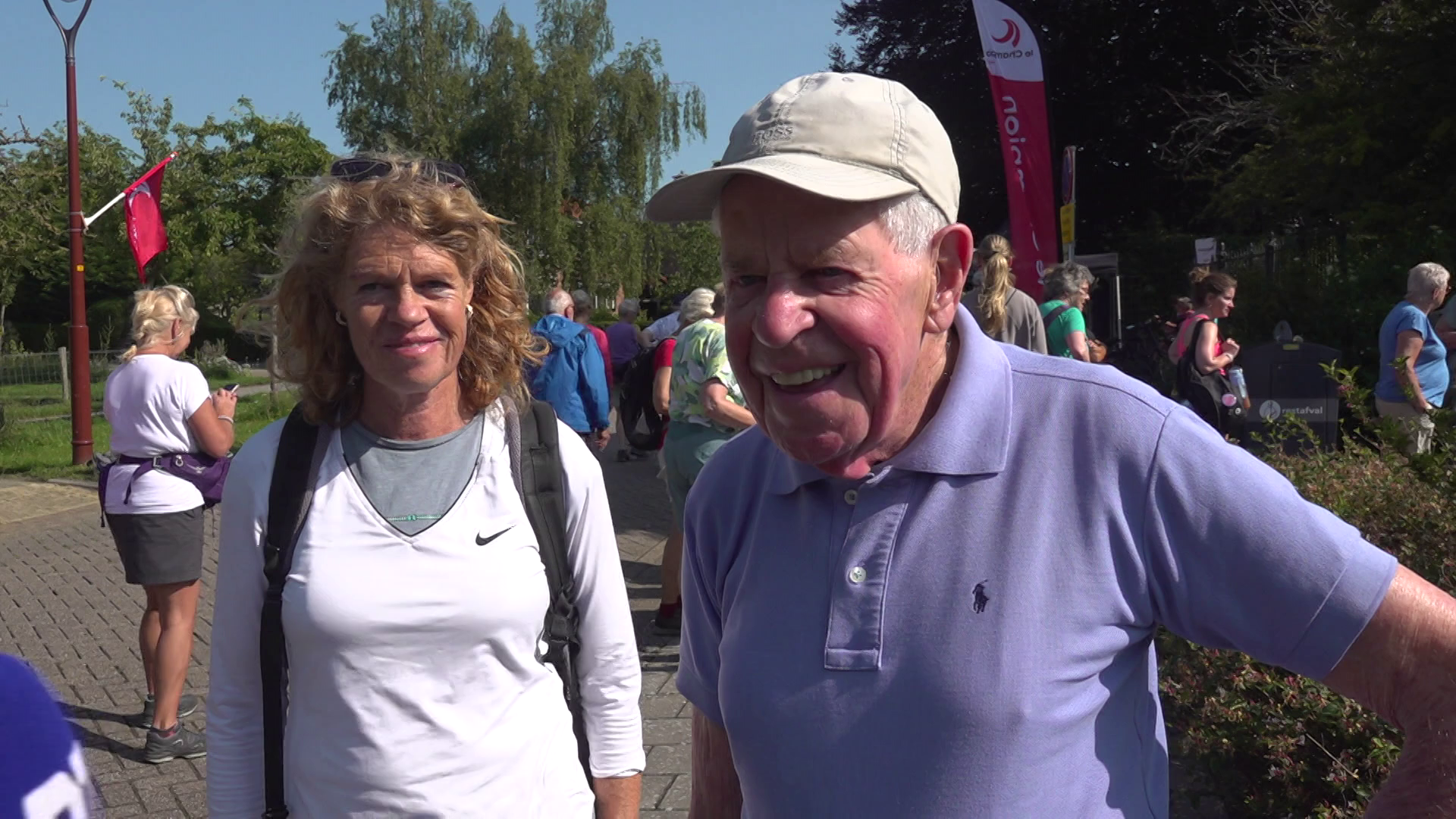 Een vrouw en een man staan buiten op een zonnige dag met een groep mensen op de achtergrond. De vrouw draagt een witte trui en een rugzak, terwijl de man een paarse polo en een pet op heeft. Ze kijken lachend naar de camera.