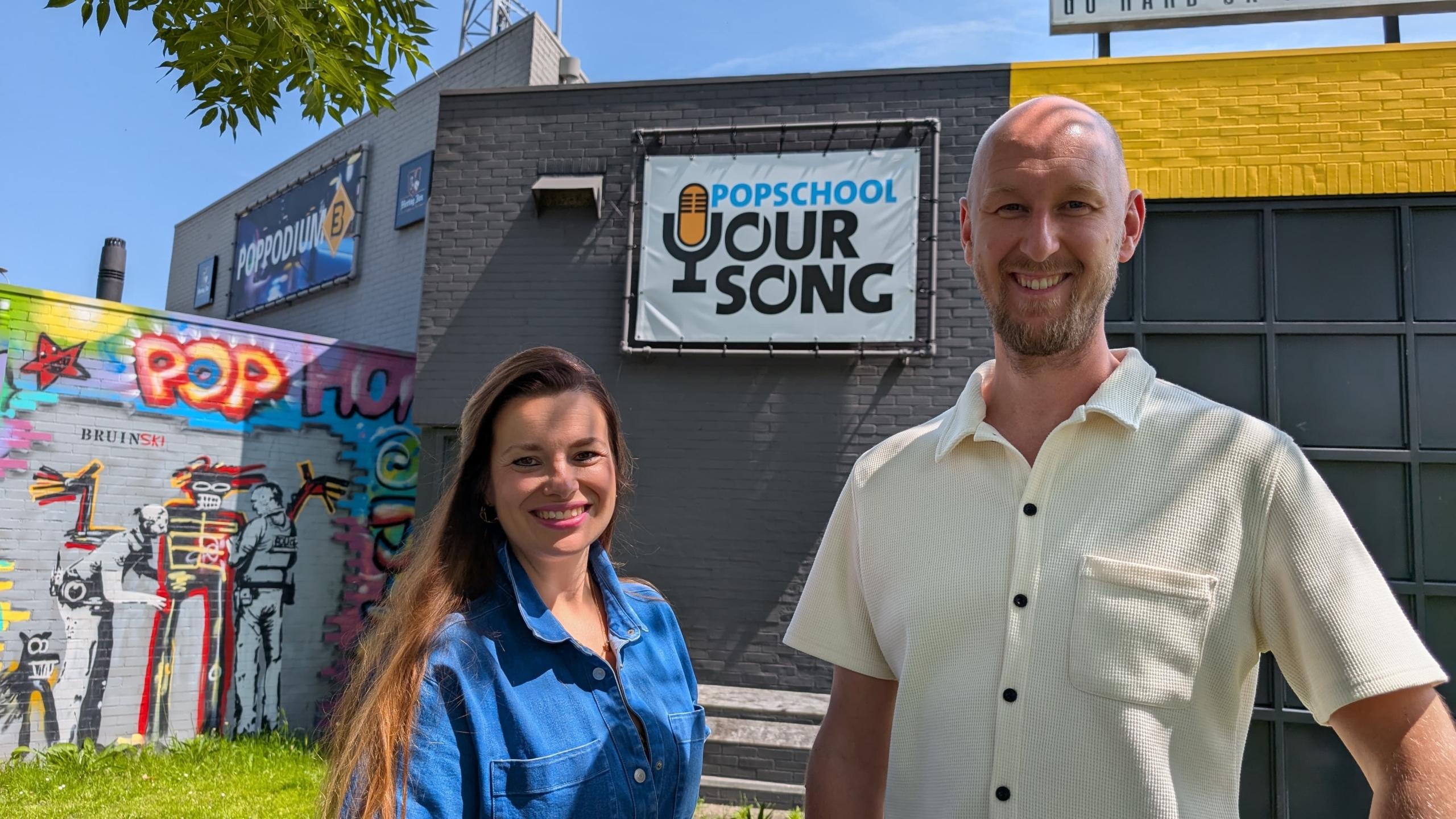 Twee mensen staan voor een gebouw met het bord 'Popschool Your Song'. De muur is versierd met kleurrijke graffiti. Het is een zonnige dag.