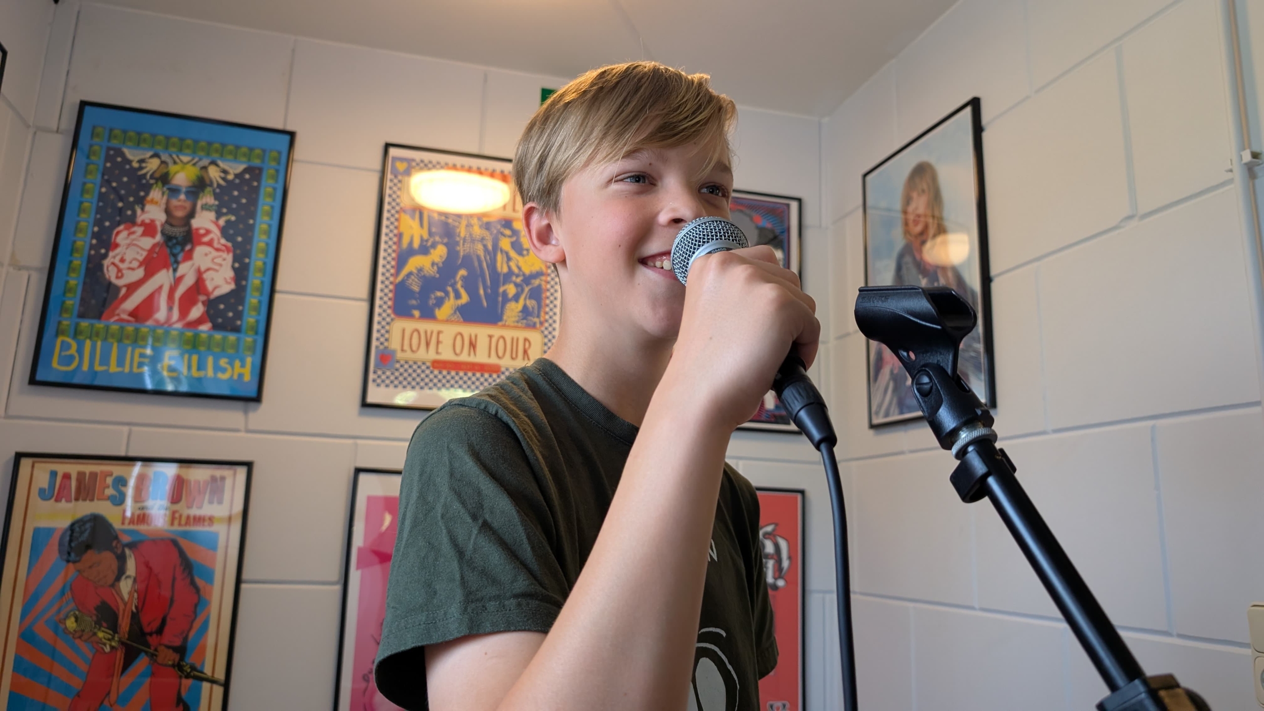 Jongen in een muziekruimte houdt een microfoon vast en zingt, omringd door posters van artiesten aan de muren.