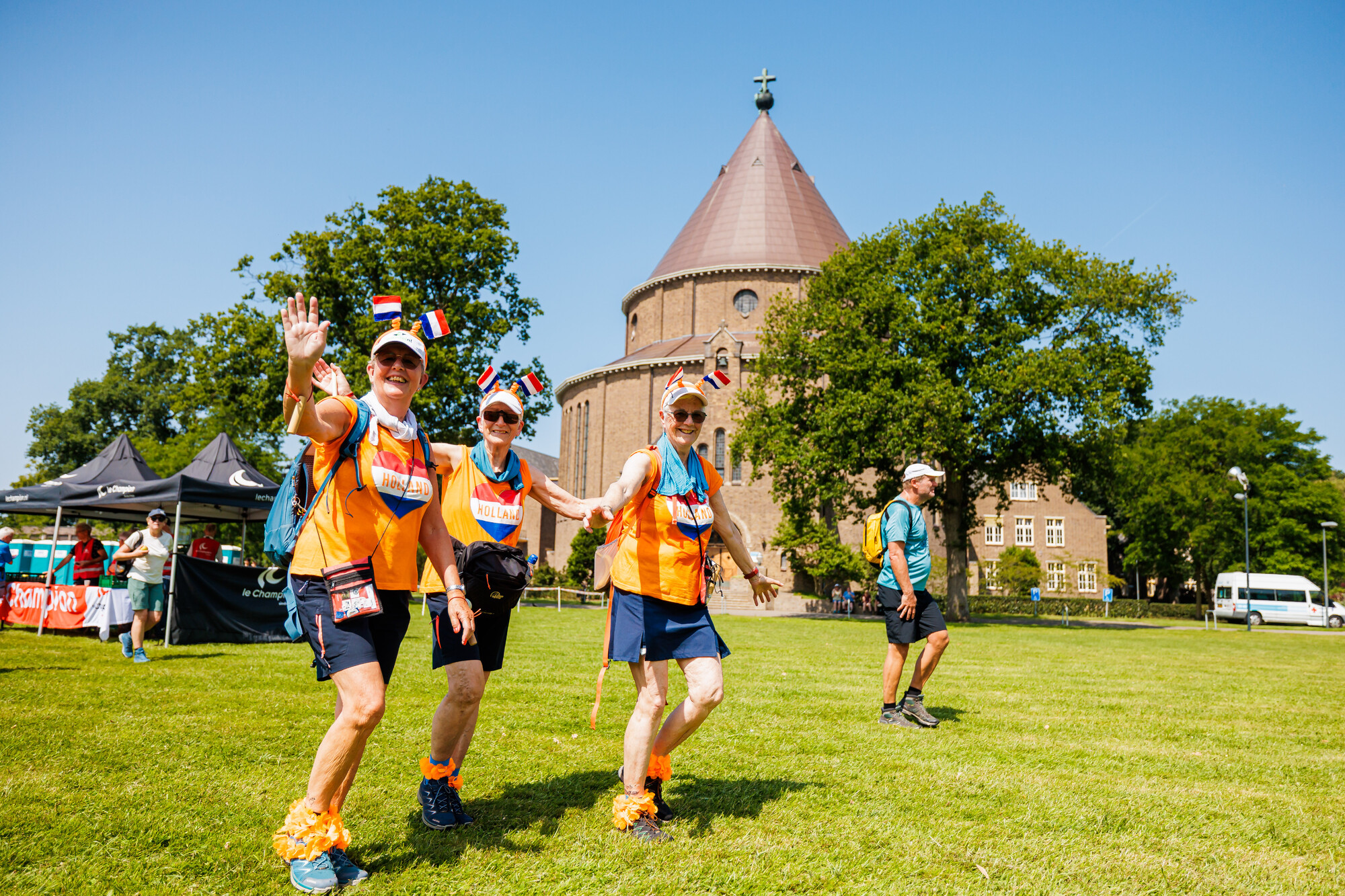 Drie mensen gekleed in oranje shirts met de tekst 'Holland', dragen Nederlandse vlagaccessoires op hun hoofd en begroeten de camera terwijl ze op een grasveld voor een rond gebouw wandelen, met een vierde persoon en festivalstands op de achtergrond.
