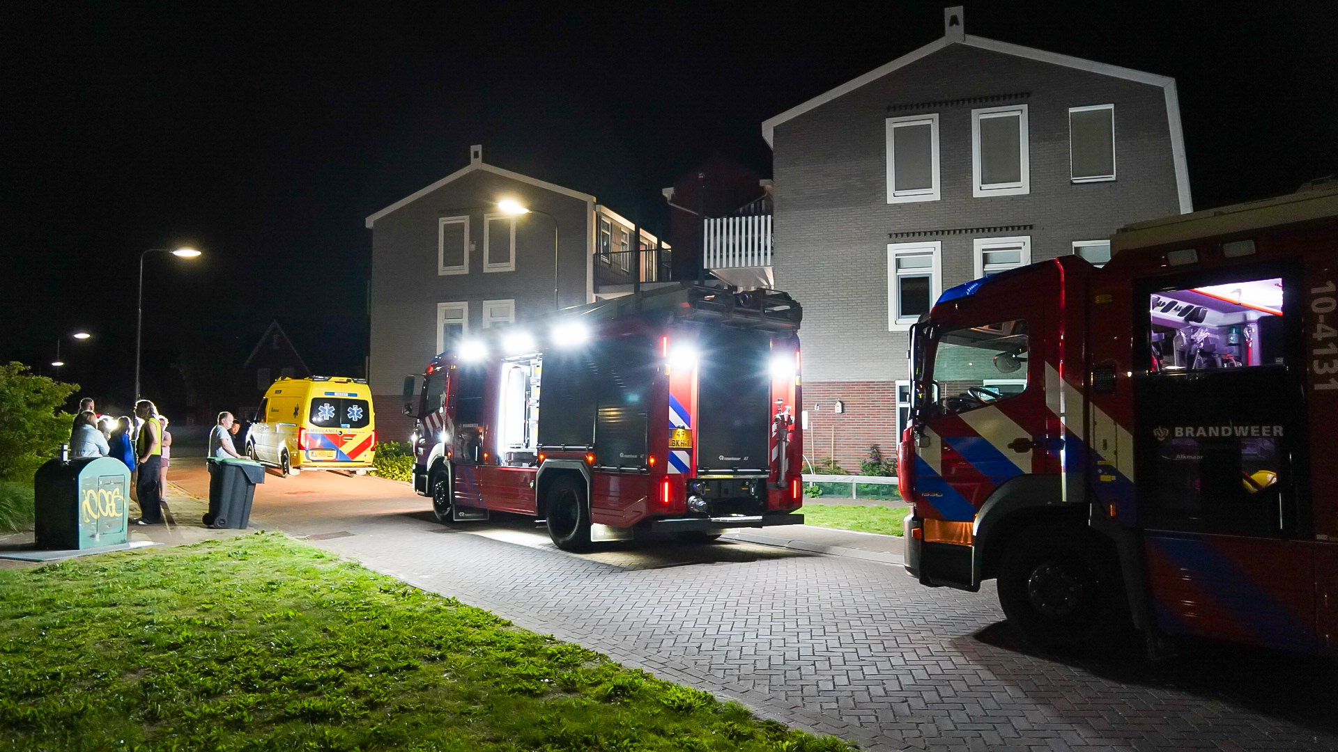 Brandweervoertuigen en een ambulance staan 's nachts geparkeerd op een straat in een woonwijk, naast een rij huizen. Mensen staan erbij en kijken naar de situatie.