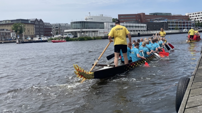 Mensen in drakenboot met turquoise shirts en rode peddels varen op een rivier, begeleid door een persoon in een geel 'CREW' shirt, met een steiger op de voorgrond en gebouwen op de achtergrond.