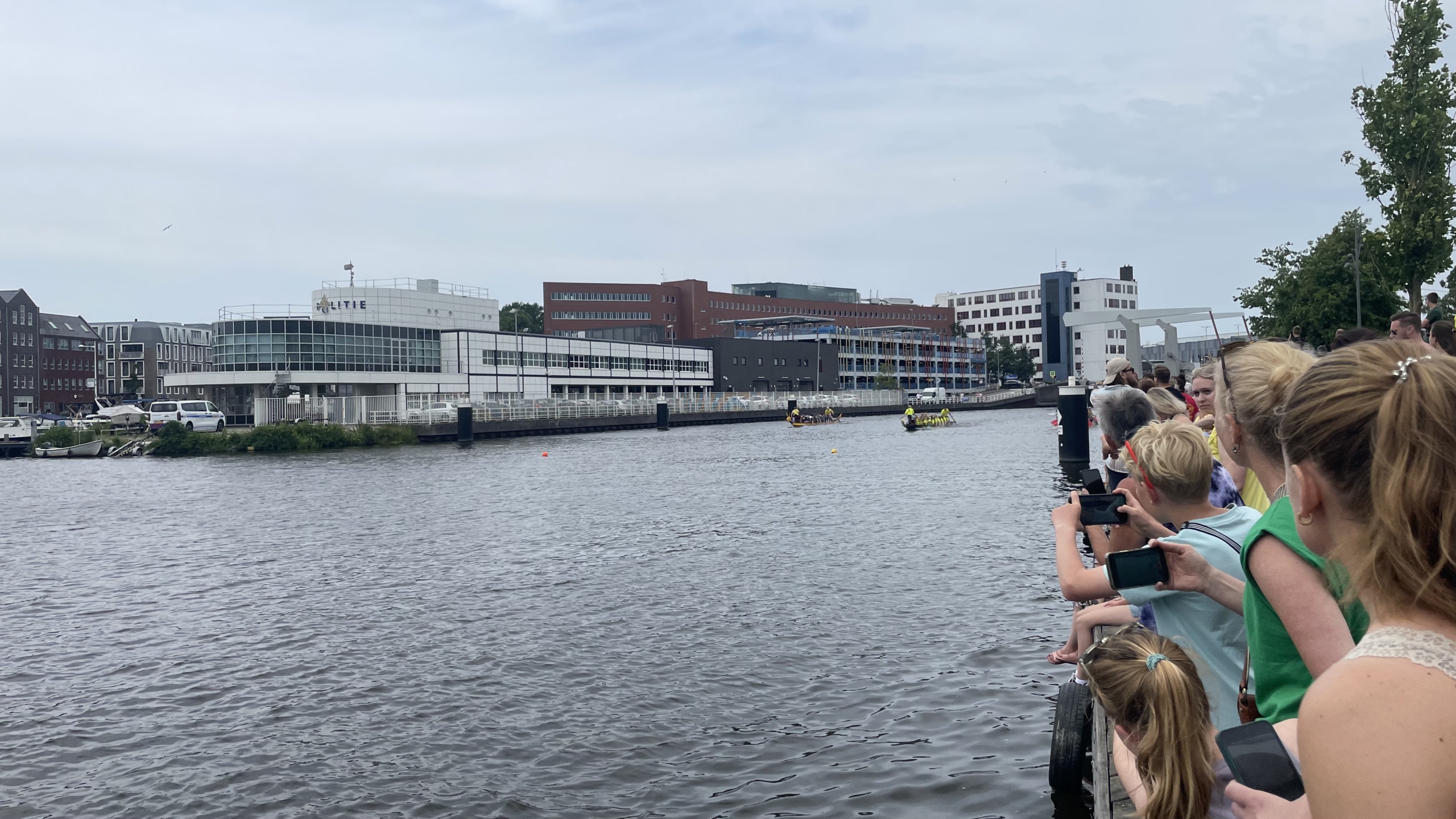 Mensen kijken uit over een rivier naar een politiebureau en een botenrace, met twee boten die parallel varen voor een rij moderne gebouwen in de achtergrond.