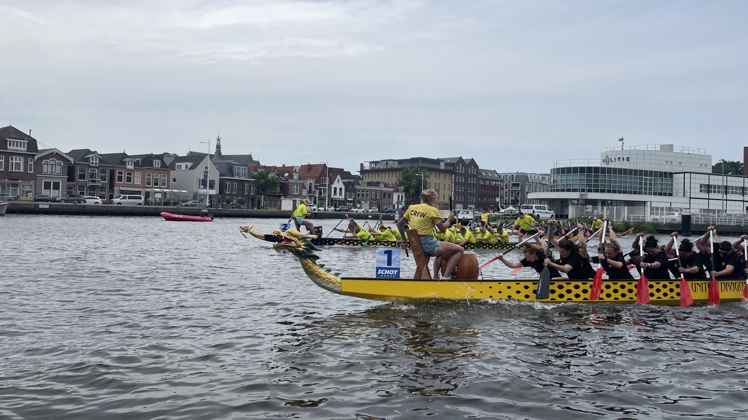 Drakenbotenrace met twee gele boten met drakenkoppen voorop, teams van mensen in gele en zwarte shirts peddelen synchroon, op een rivier met stadsgebouwen op de achtergrond.