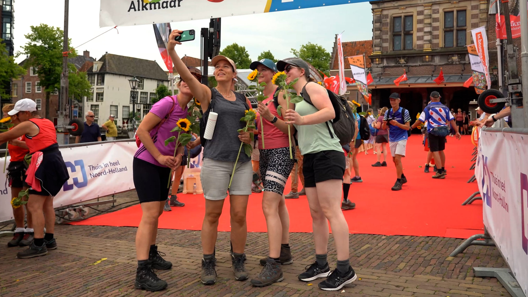 Vier wandelaars poseren voor een selfie bij de finishlijn van de Wandel4daagse in Alkmaar, elk met een zonnebloem in de hand, op een rode loper met toeschouwers op de achtergrond.