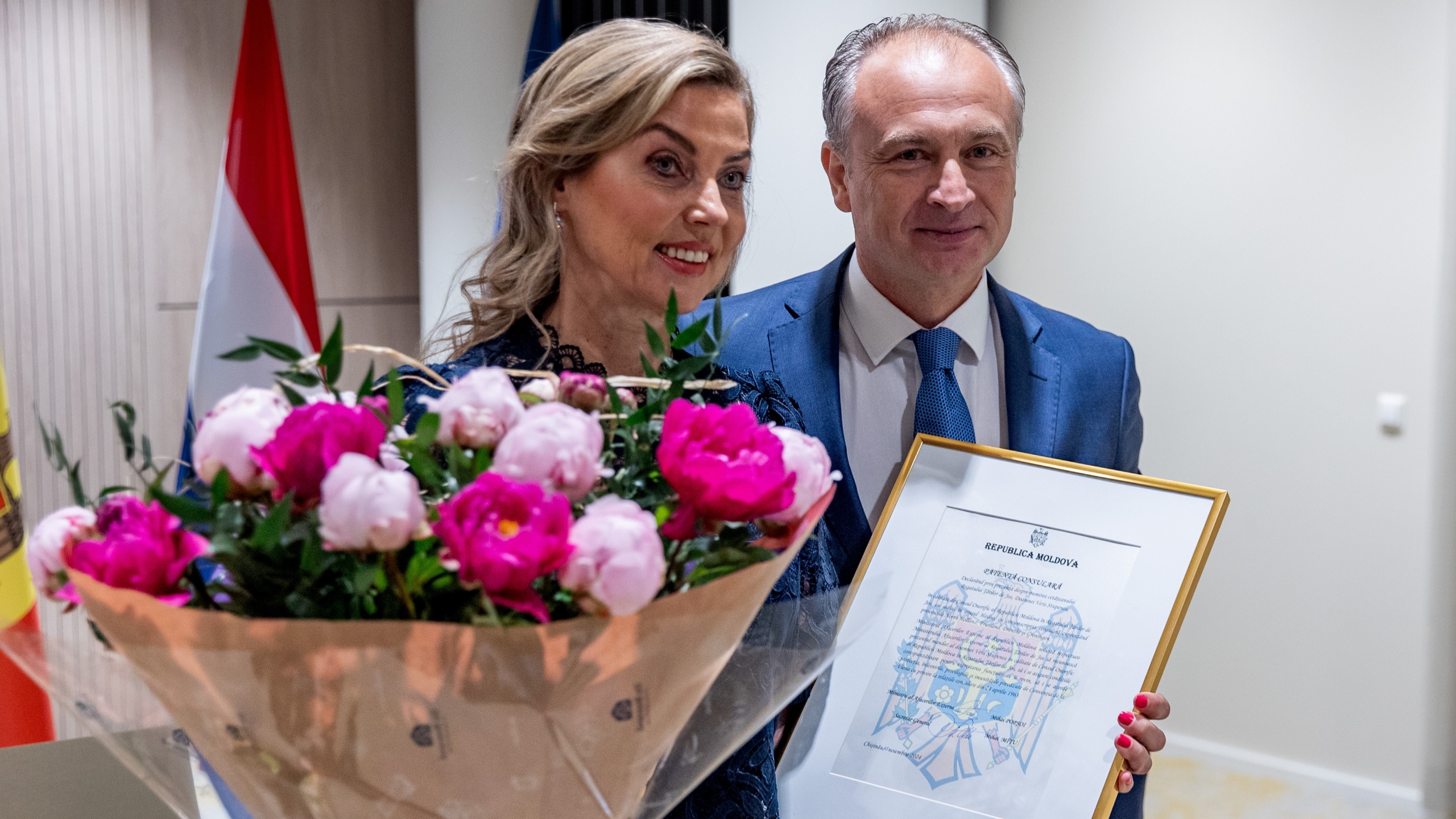Een man en een vrouw poseren samen; de vrouw houdt een groot boeket roze bloemen vast, terwijl de man een ingelijste oorkonde toont. Achter hen zijn de vlaggen van Moldavië en een ander land zichtbaar.