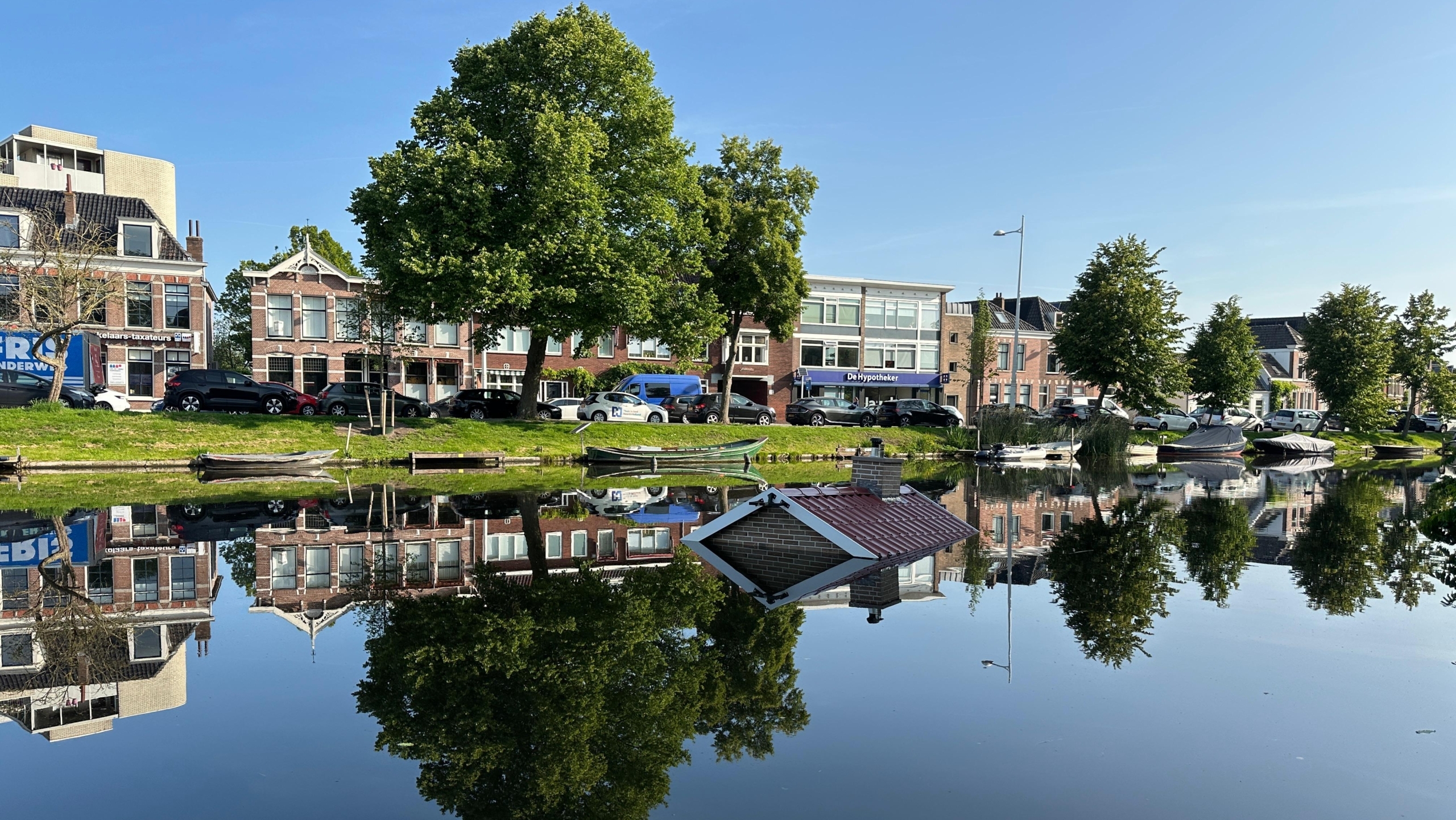 Gracht met huizen en bomen erlangs, met een dak van een huis dat onder water lijkt te staan, alles weerspiegeld in het kalme wateroppervlak.