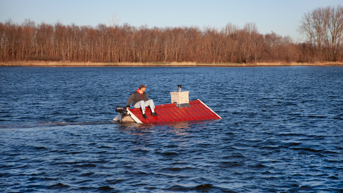 Een persoon zit op een schuins dak dat gedeeltelijk onder water is, met een motor eraan bevestigd, op een meer met een bebost gebied op de achtergrond."