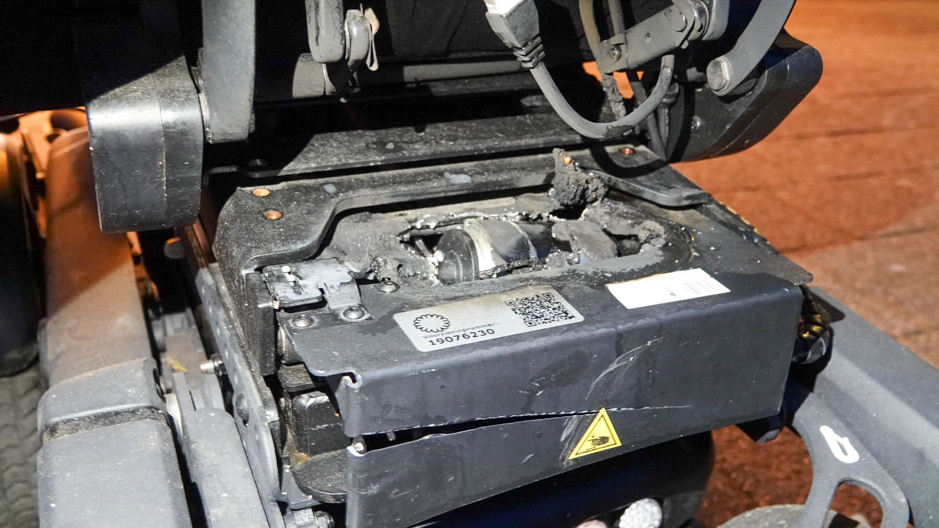 Een beschadigd onderdeel van een machine met zichtbare brandplekken en roet, waarbij een sticker met een QR-code en serienummer zichtbaar is.