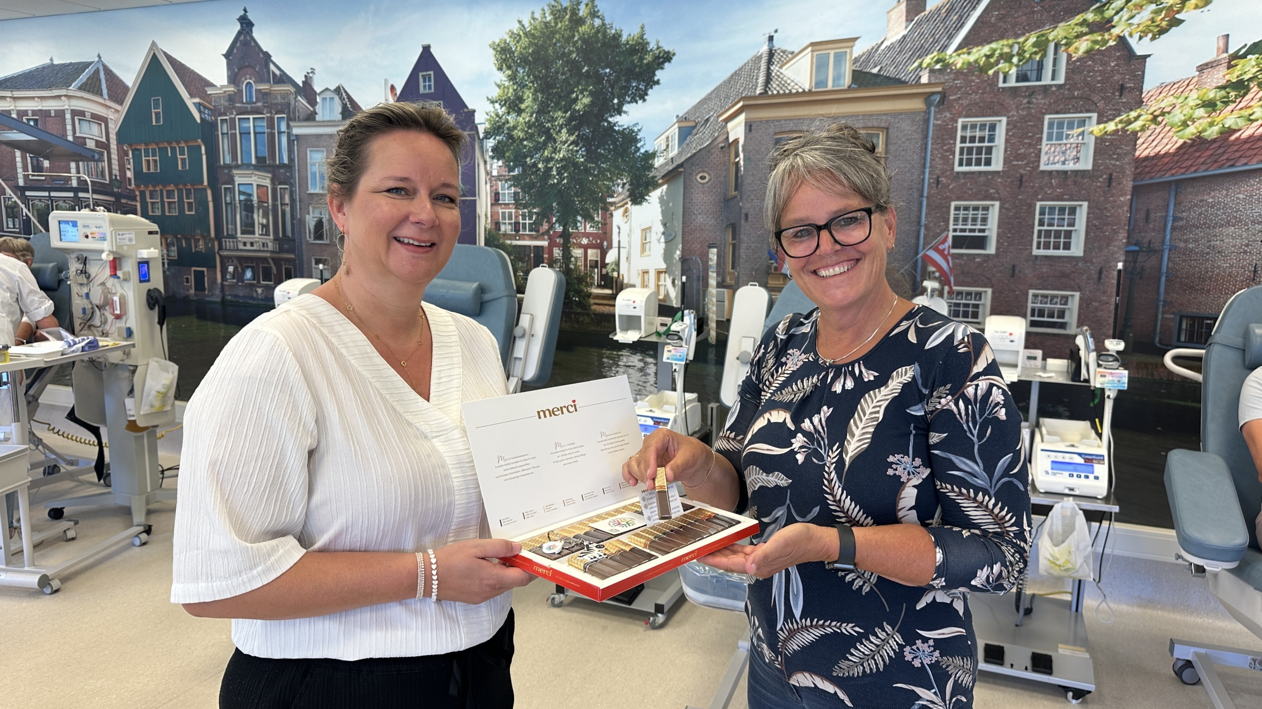 Twee vrouwen staan glimlachend in een ruimte met donatie-apparatuur, terwijl ze een Merci chocolade doos vasthouden; de achtergrond toont een grote foto van traditionele Nederlandse gebouwen.