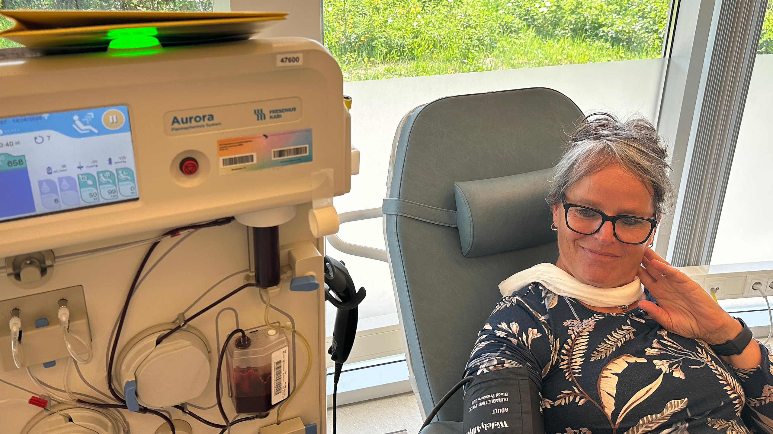 Vrouw in een stoel ondergaat een medische procedure met een machine links in beeld, binnen een ruimte met ramen en uitzicht op gras en bomen.