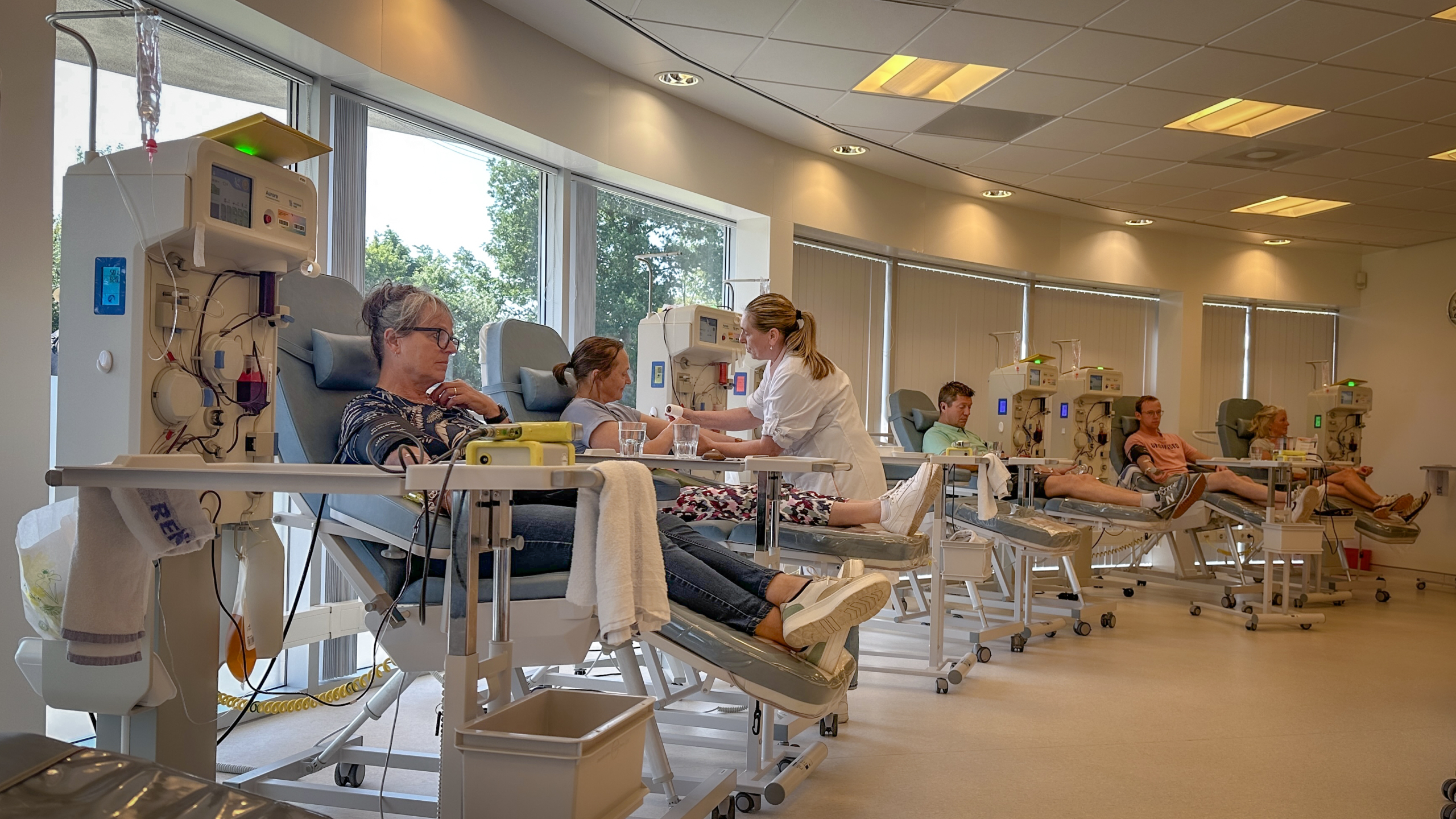 Mensen die in een kliniek bloeddonatie of dialyse ondergaan, zitten in stoelen naast medische apparaten. Een verpleegkundige helpt een van de patiënten. De ruimte is helder verlicht met grote ramen aan de zijkant.