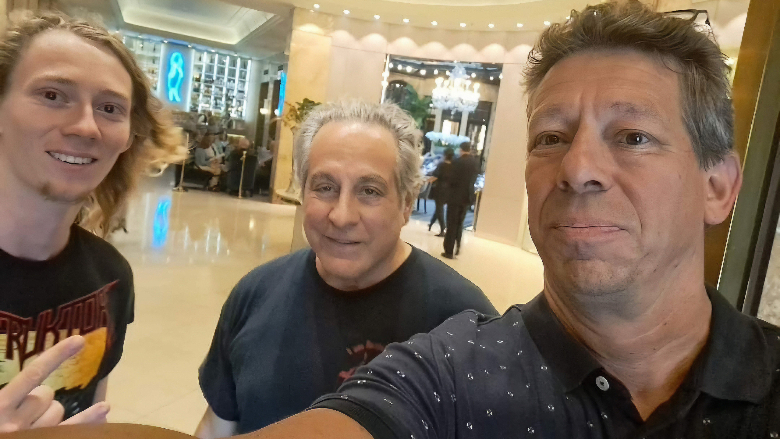 Drie mannen poseren voor een selfie in een verlichte lobby met een kroonluchter op de achtergrond.
