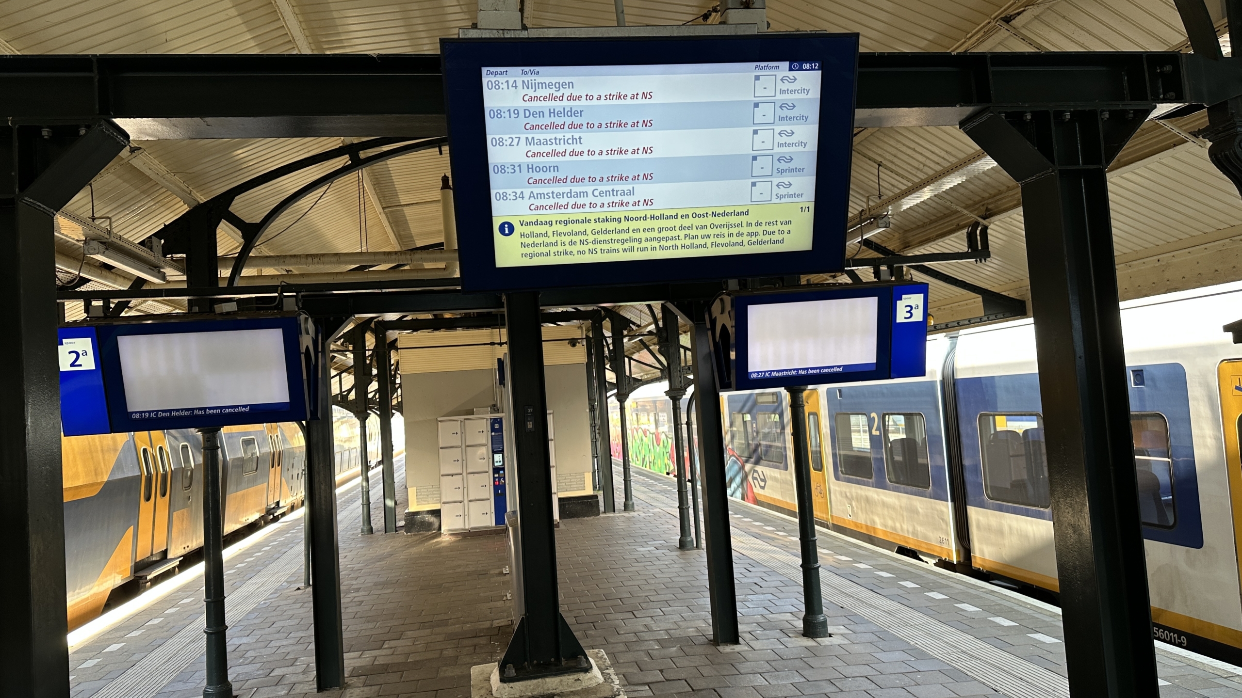 Een leeg treinstation met een NS-trein op de achtergrond en meerdere informatieschermen met berichten over treinannuleringen vanwege een staking.