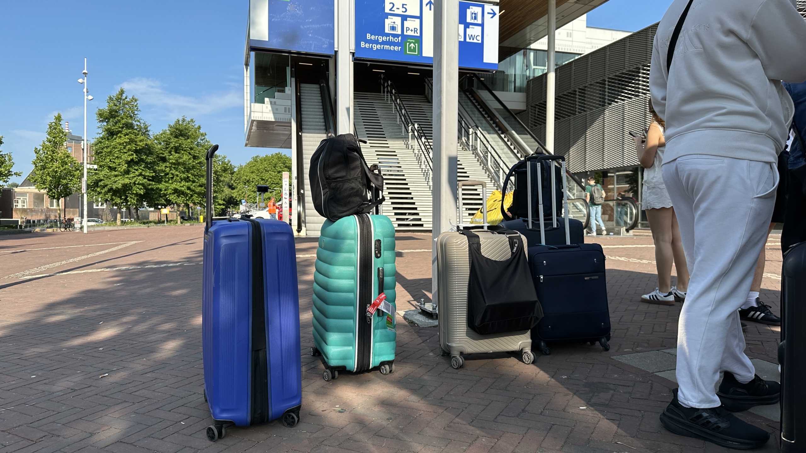 Koffers en mensen bij een station met bewegwijzering naar verschillende perrons en voorzieningen, op een zonnige dag.