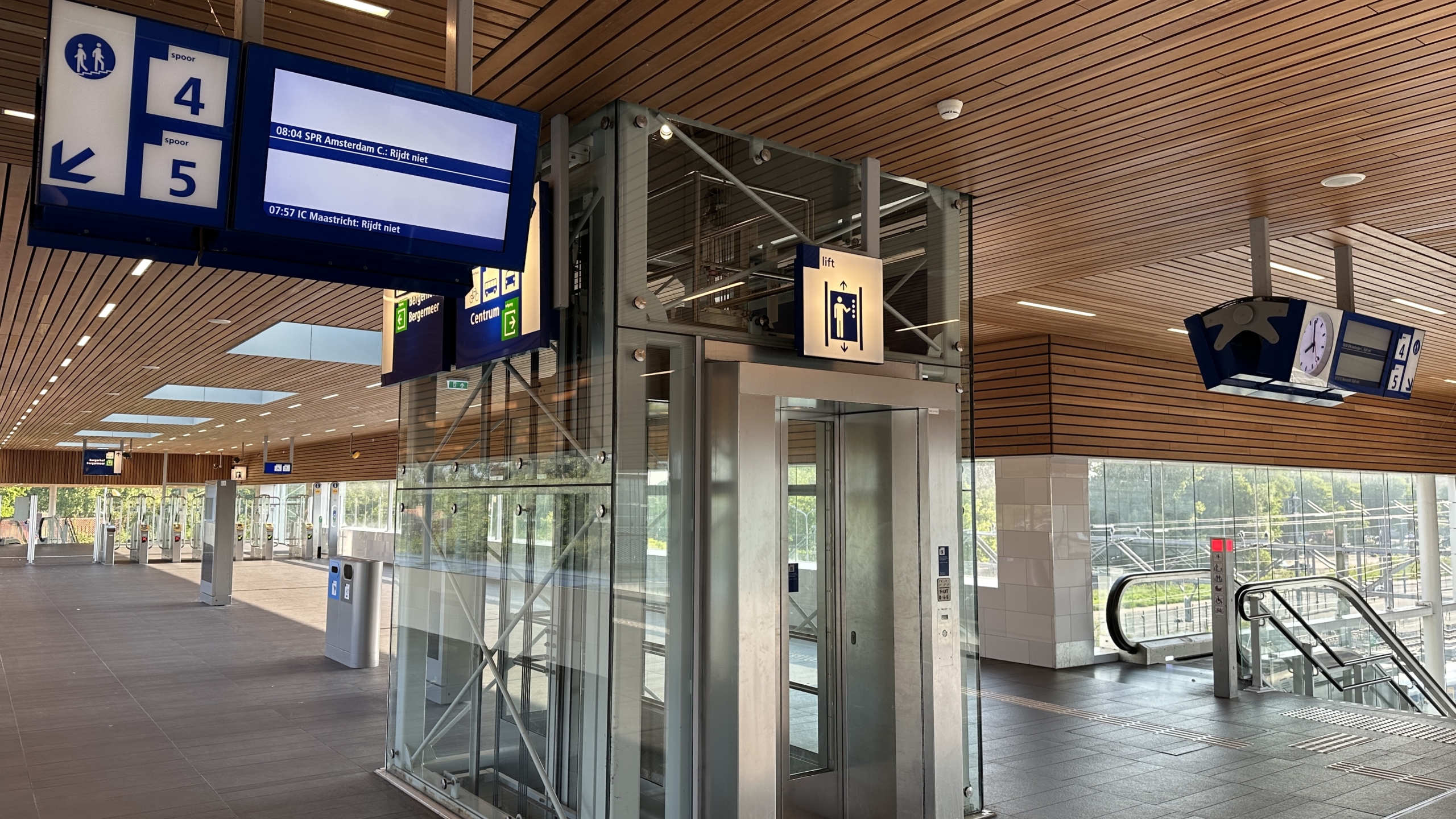 Modern treinstation met een glazen lift, richtingbordjes voor perrons 4 en 5, en een roltrap. Er zijn digitale schermen met treininformatie en een klok in een interieur met houten plafond.