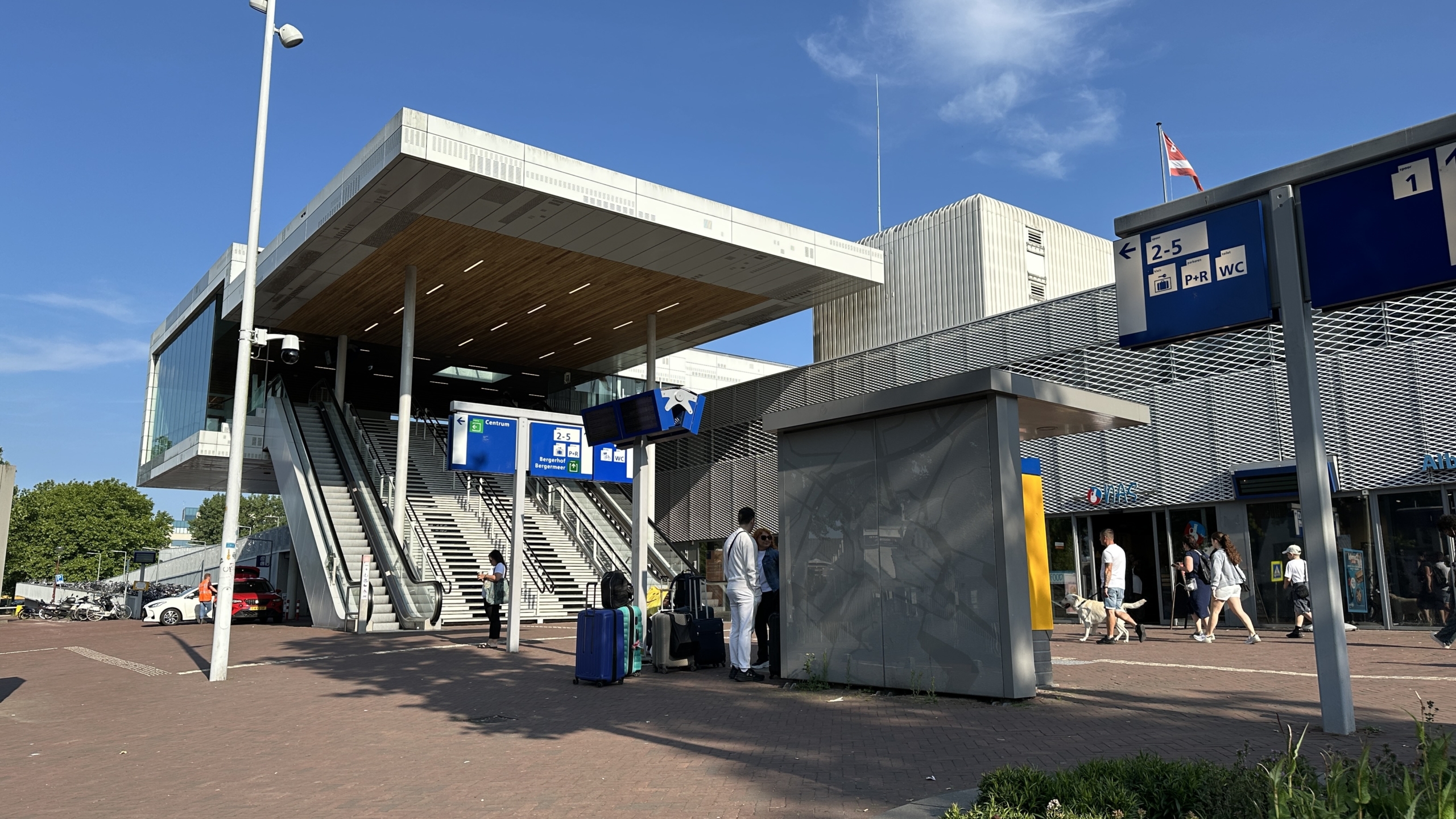 Buitenkant van een trein- of busstation met een brede trap en roltrap naar de ingang, omringd door mensen met bagage. Borden wijzen routes aan en er is helder weer met blauwe lucht.