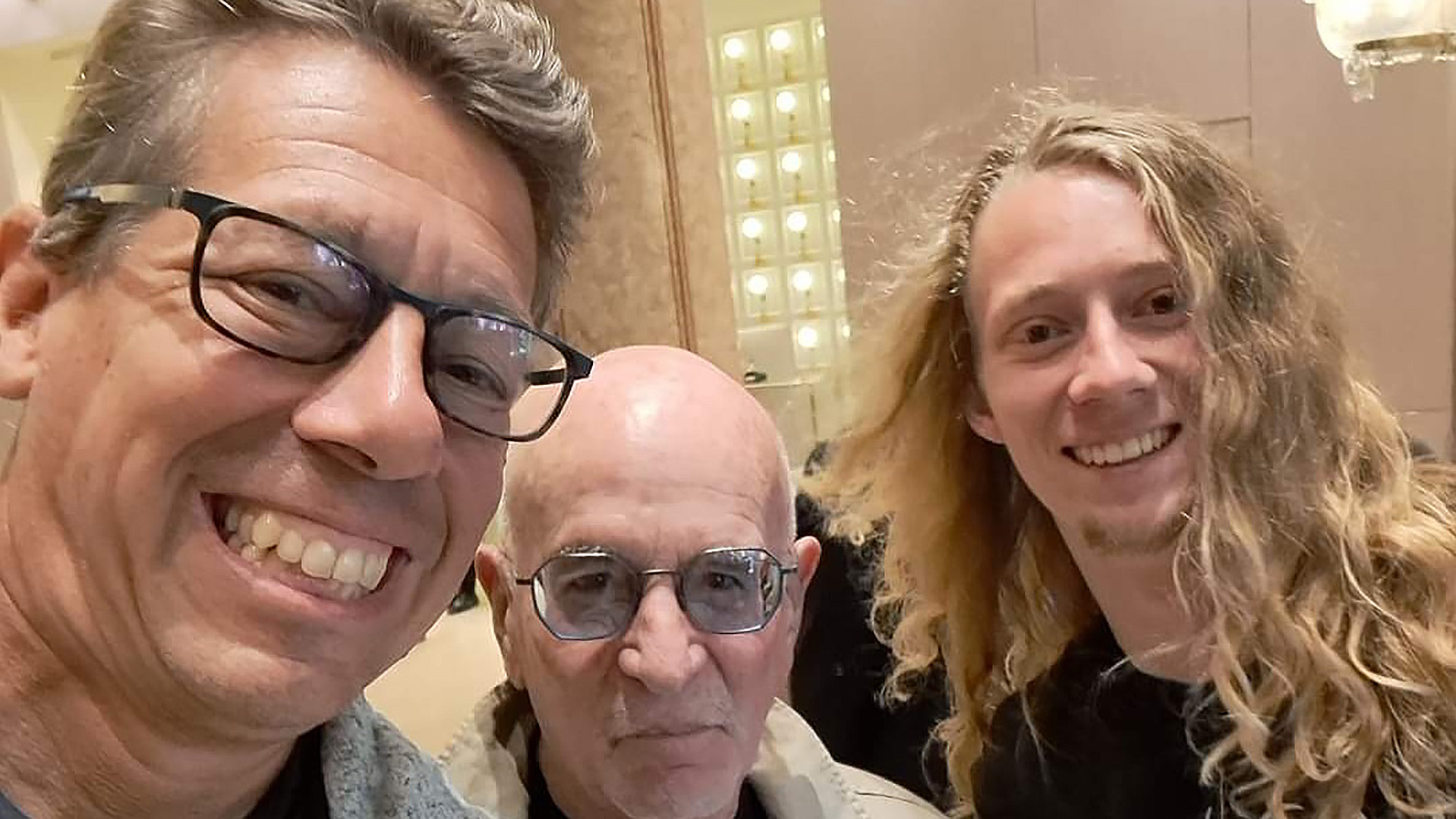 Drie mannen poseren voor een selfie, twee glimlachend en een met een neutraal gezicht, in een binnenruimte met warme verlichting.