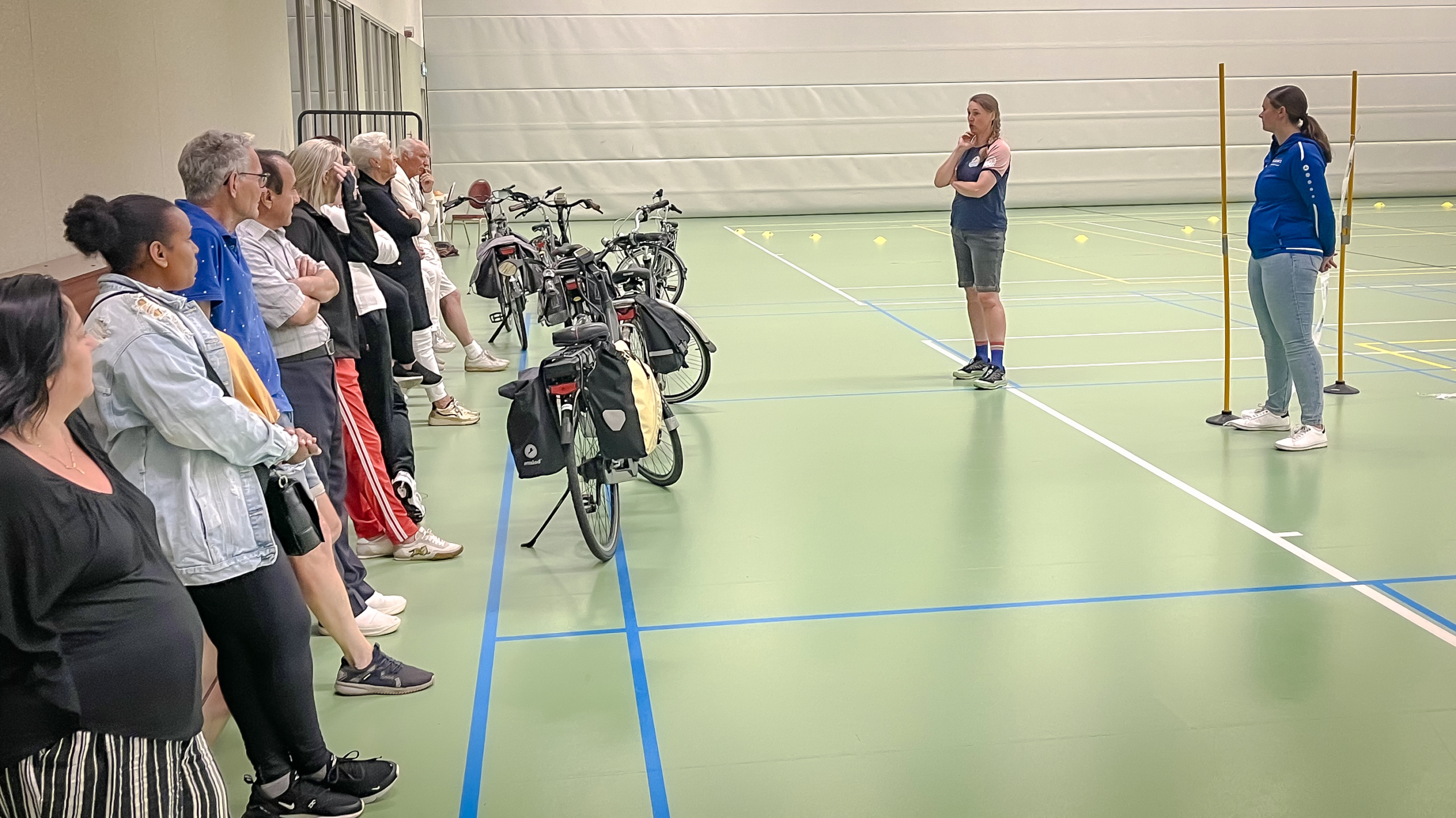 Een groep mensen in een sporthal kijkt naar twee instructeurs die uitleg geven; fietsen staan in een rij naast hen.