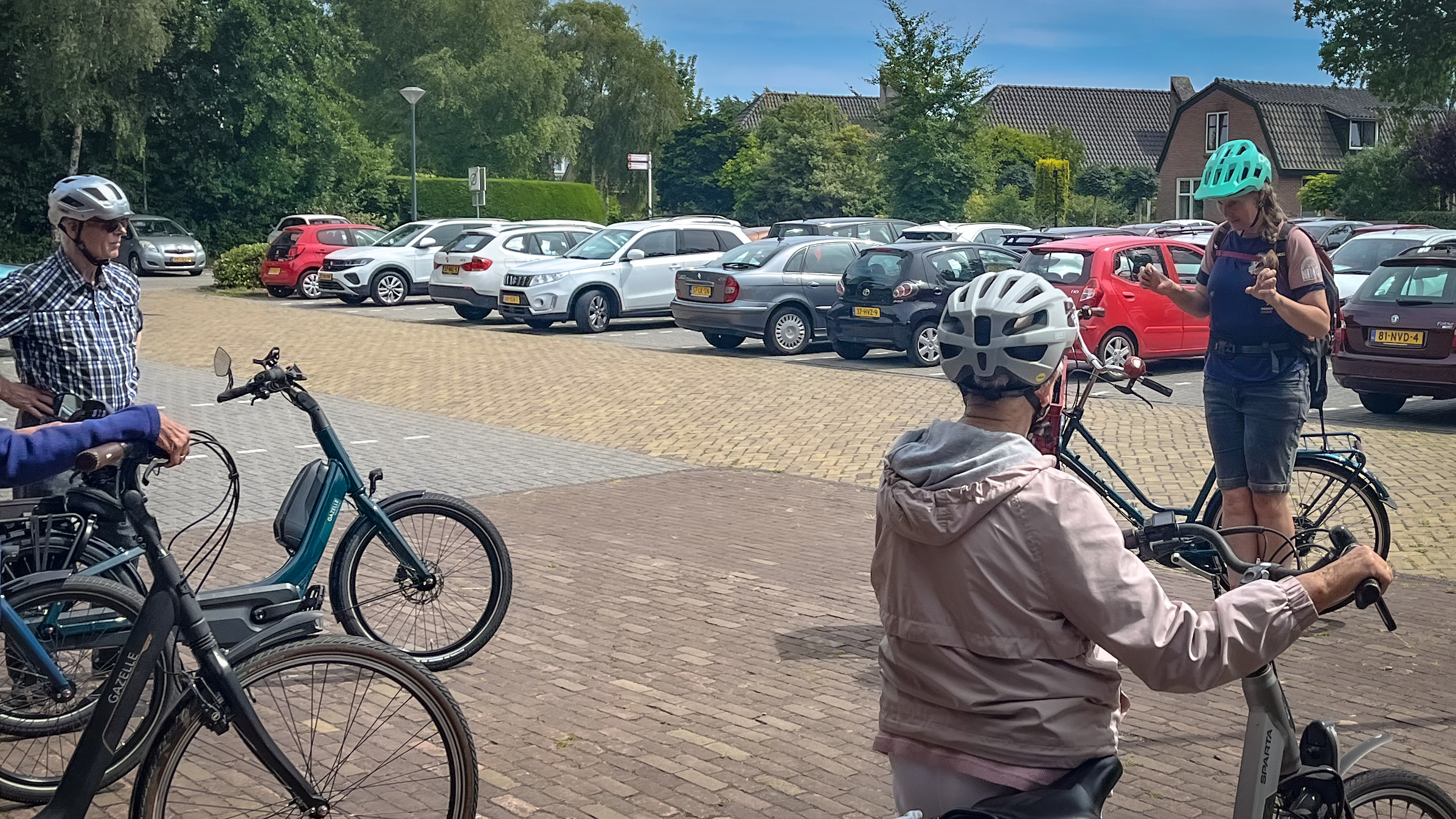 Een groep mensen met fietsen en helmen staat op een parkeerplaats met auto's op de achtergrond bij daglicht.