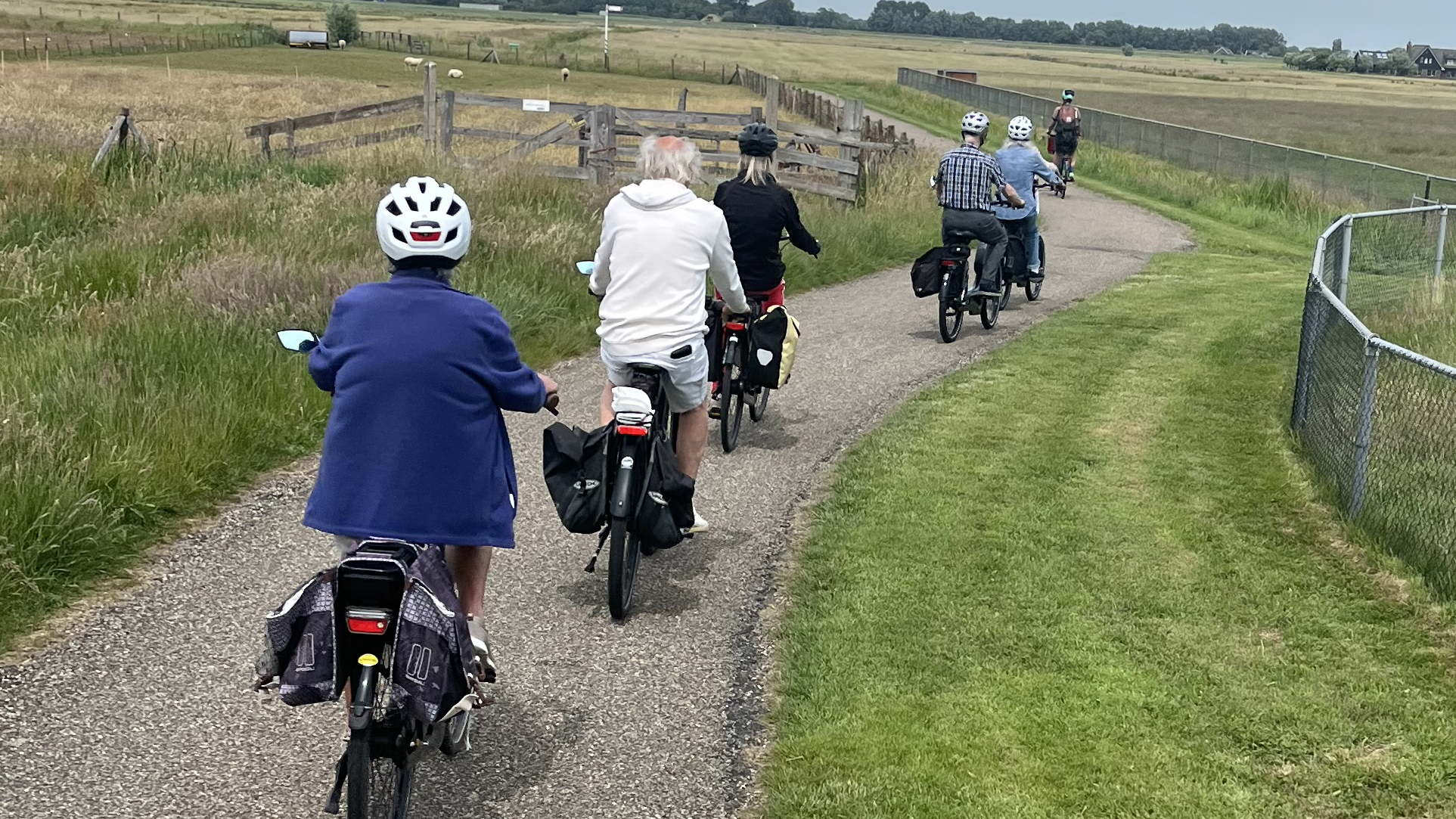 Een groep mensen fietst op een smal pad door een landelijk gebied, omgeven door grasvelden en landbouwgrond.