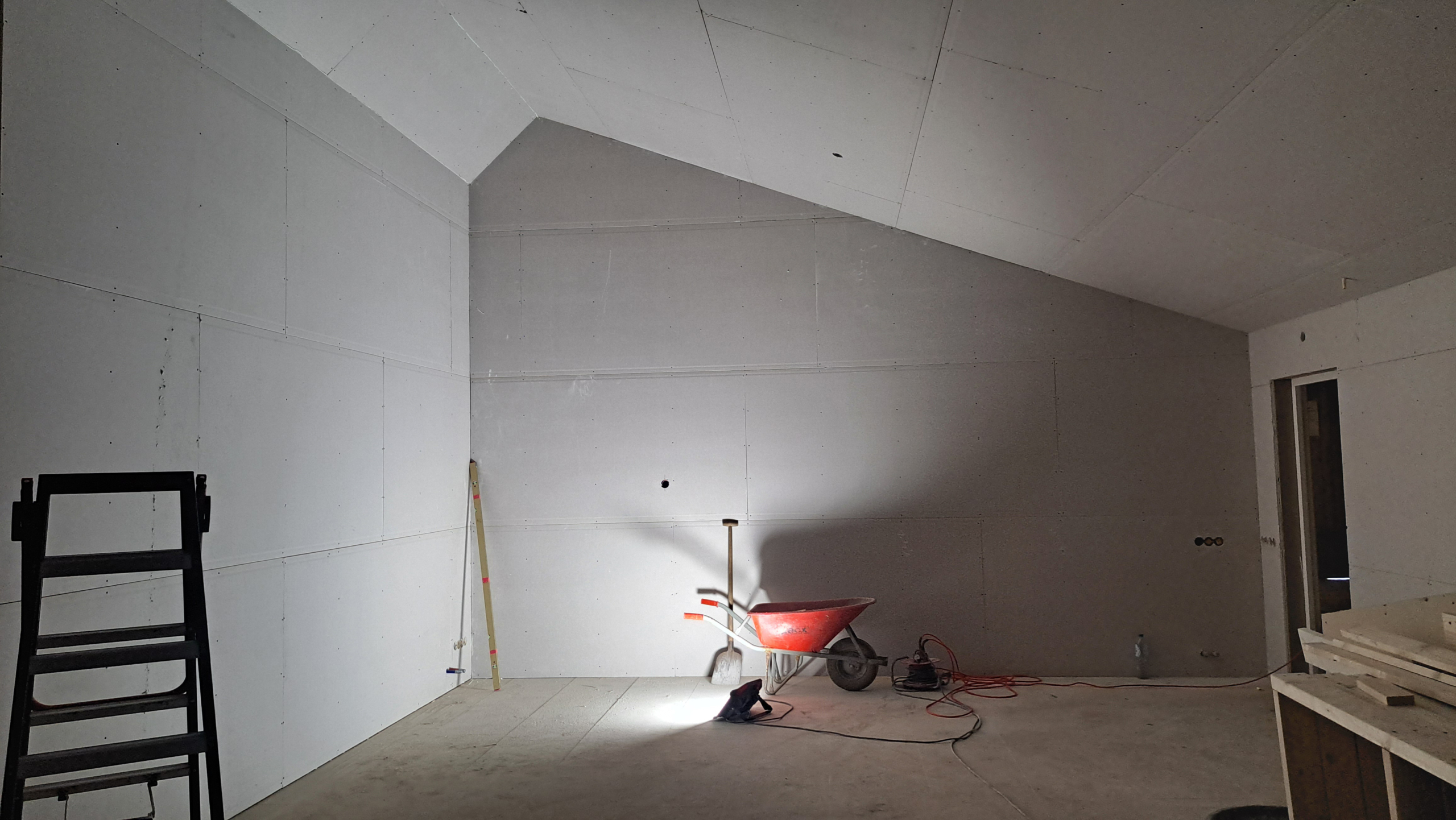 Een lege kamer in aanbouw met gipsplaten op de muren en een schuin plafond. Er staat een rode kruiwagen en een schop in het midden, met een bouwlamp ernaast. Links is een ladder zichtbaar, en rechts zijn wat houten constructies en elektriciteitskabels te zien.