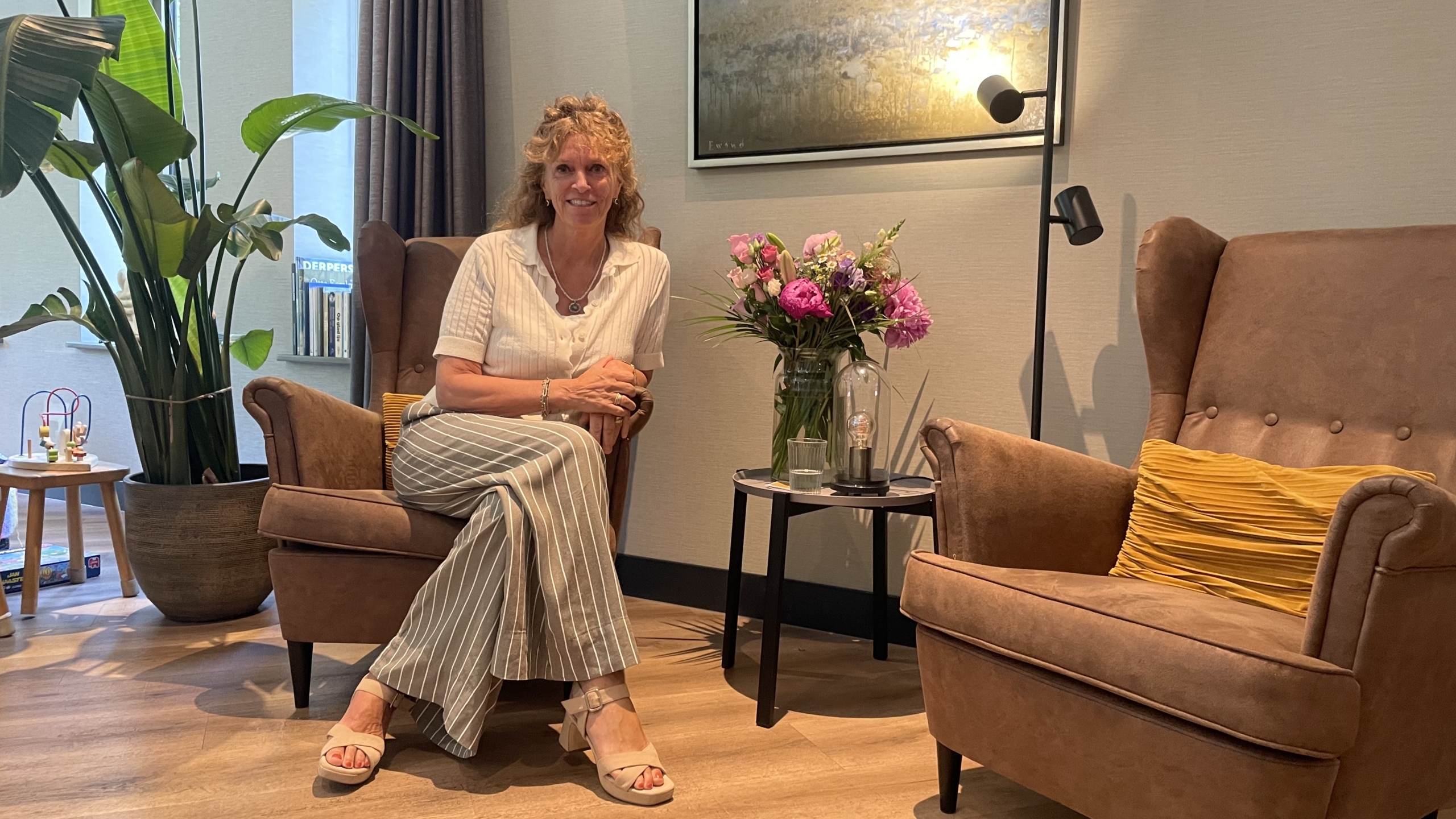 Een vrouw zit op een bruine stoel in een gezellige woonkamer, met bloemen op een tafeltje naast haar en een grote plant op de achtergrond.