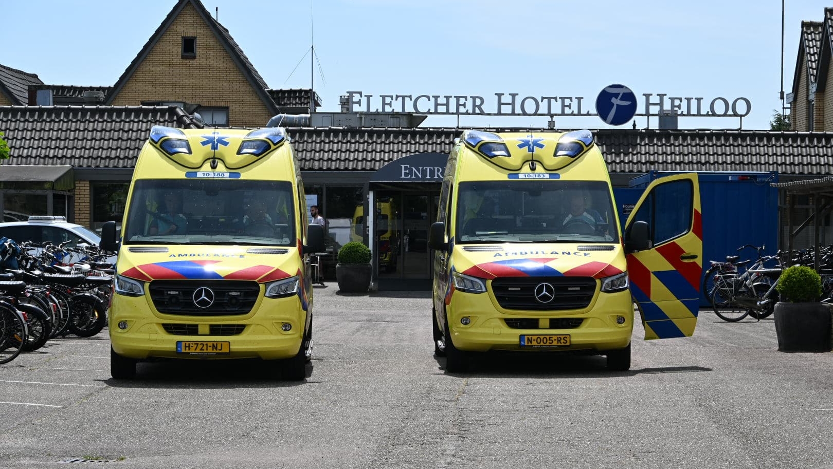 Twee gele ambulances geparkeerd voor Fletcher Hotel Heiloo, met een van de deuren van de rechterambulance open, en fietsen aan de zijkant van de parkeerplaats.