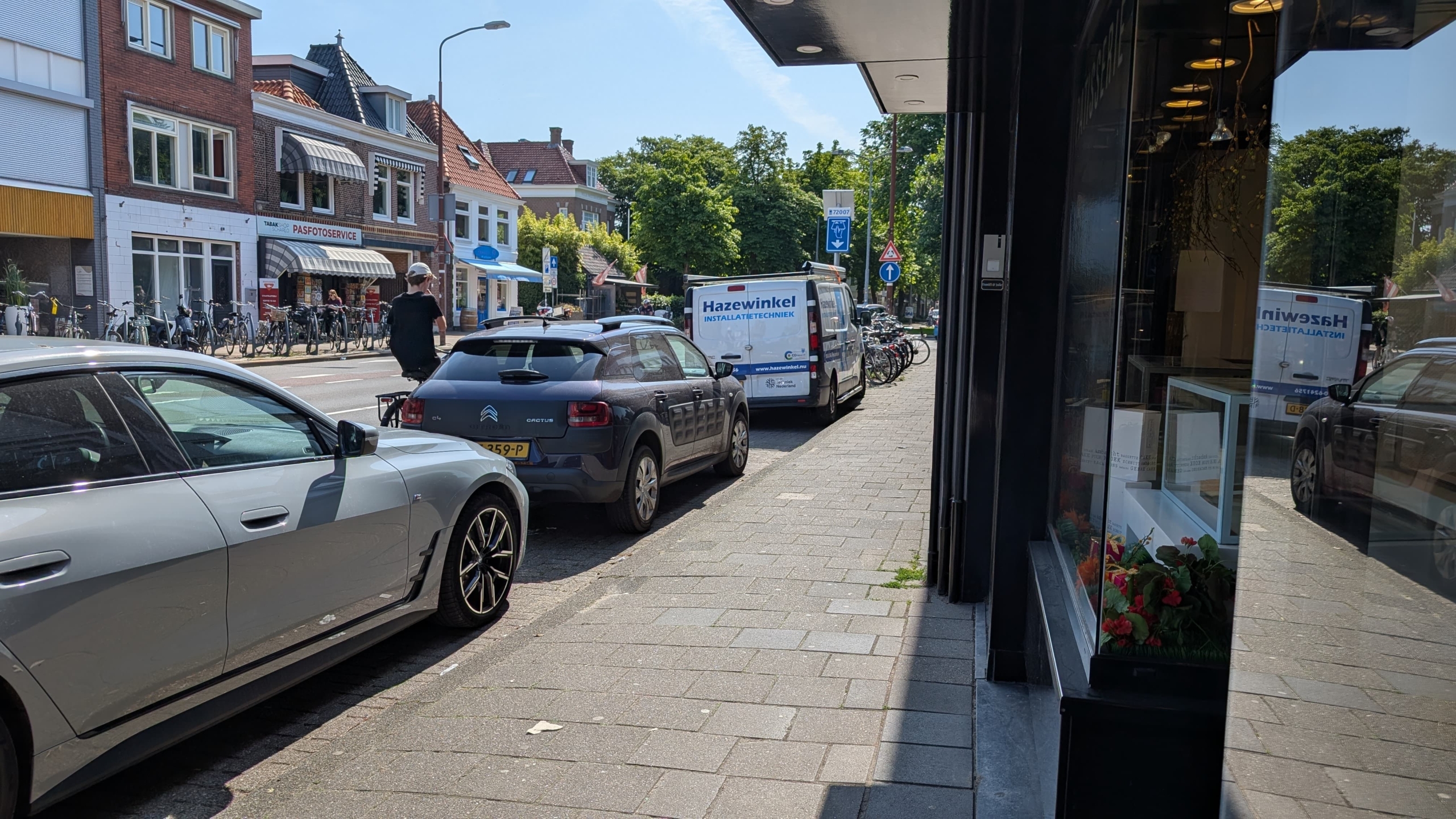 Straatbeeld met geparkeerde auto's langs de stoep en een rij met fietsen, naast enkele winkels, waaronder een pasfotoservice.