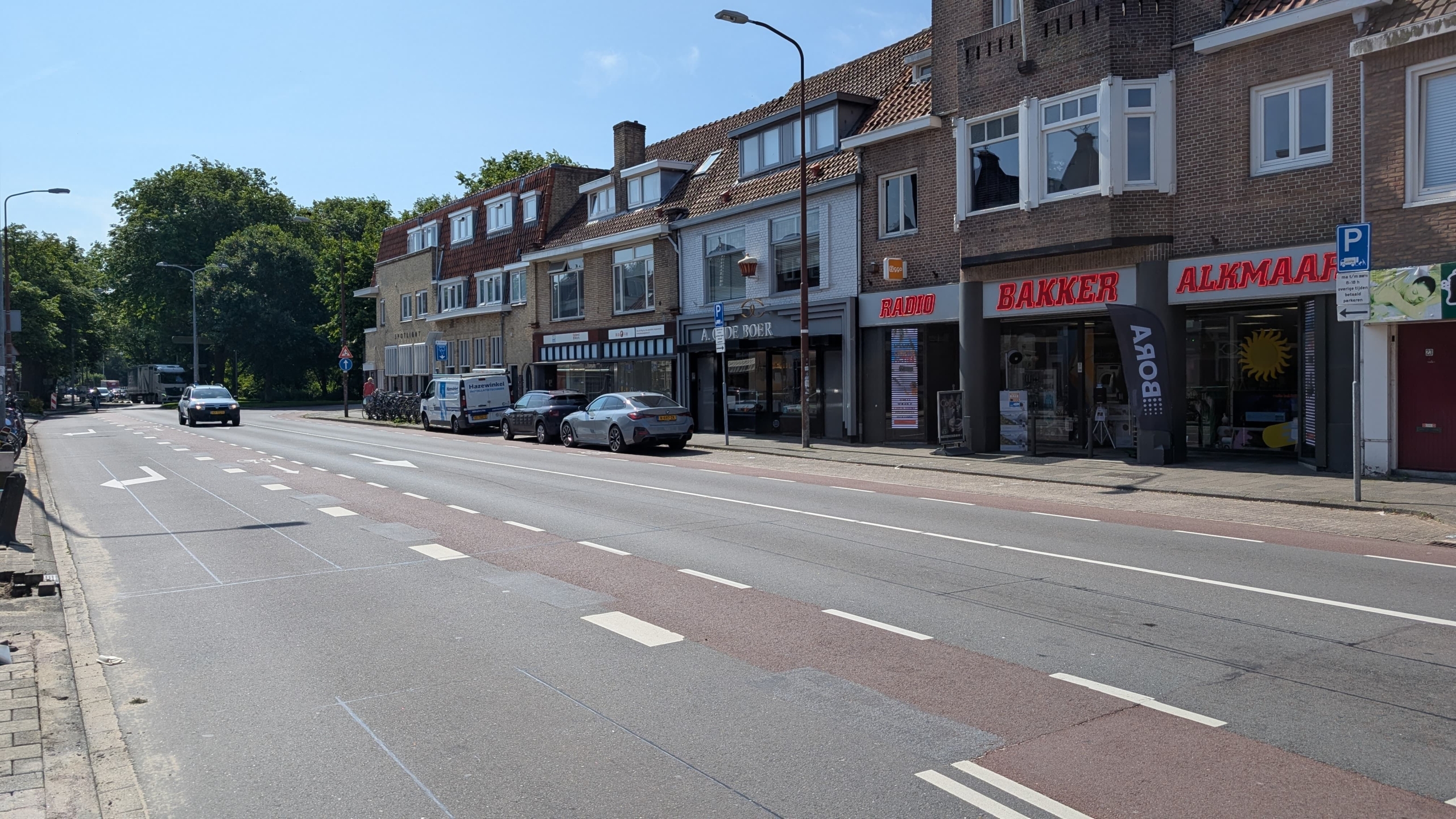 Straat met geparkeerde auto's langs een rij winkels, waaronder een bakker en een radiowinkel, onder een blauwe lucht op een zonnige dag.