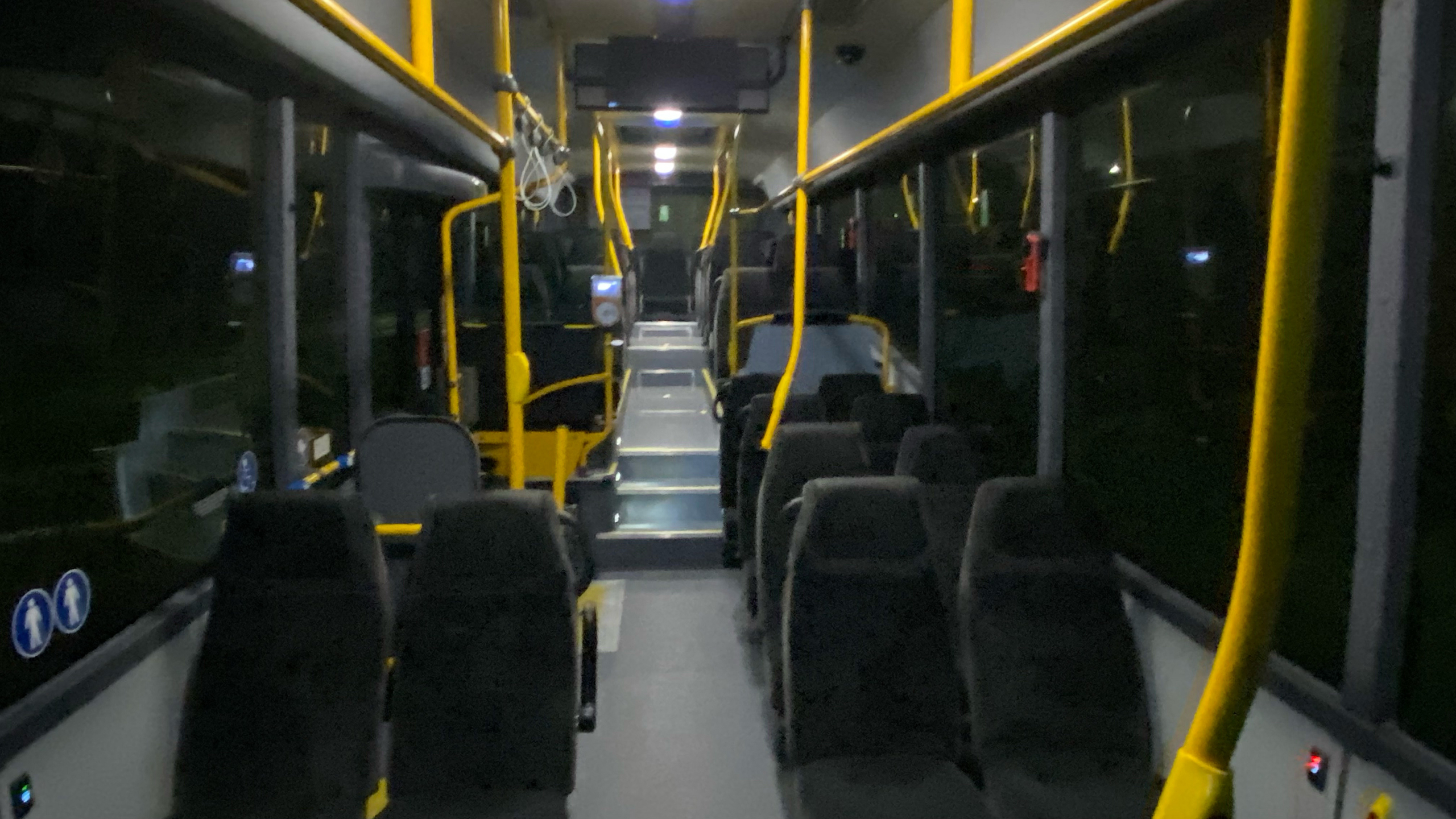 Het interieur van een lege stadsbus met zwart-grijze stoelen, gele relingen en een donkere buitenomgeving zichtbaar door de ramen.
