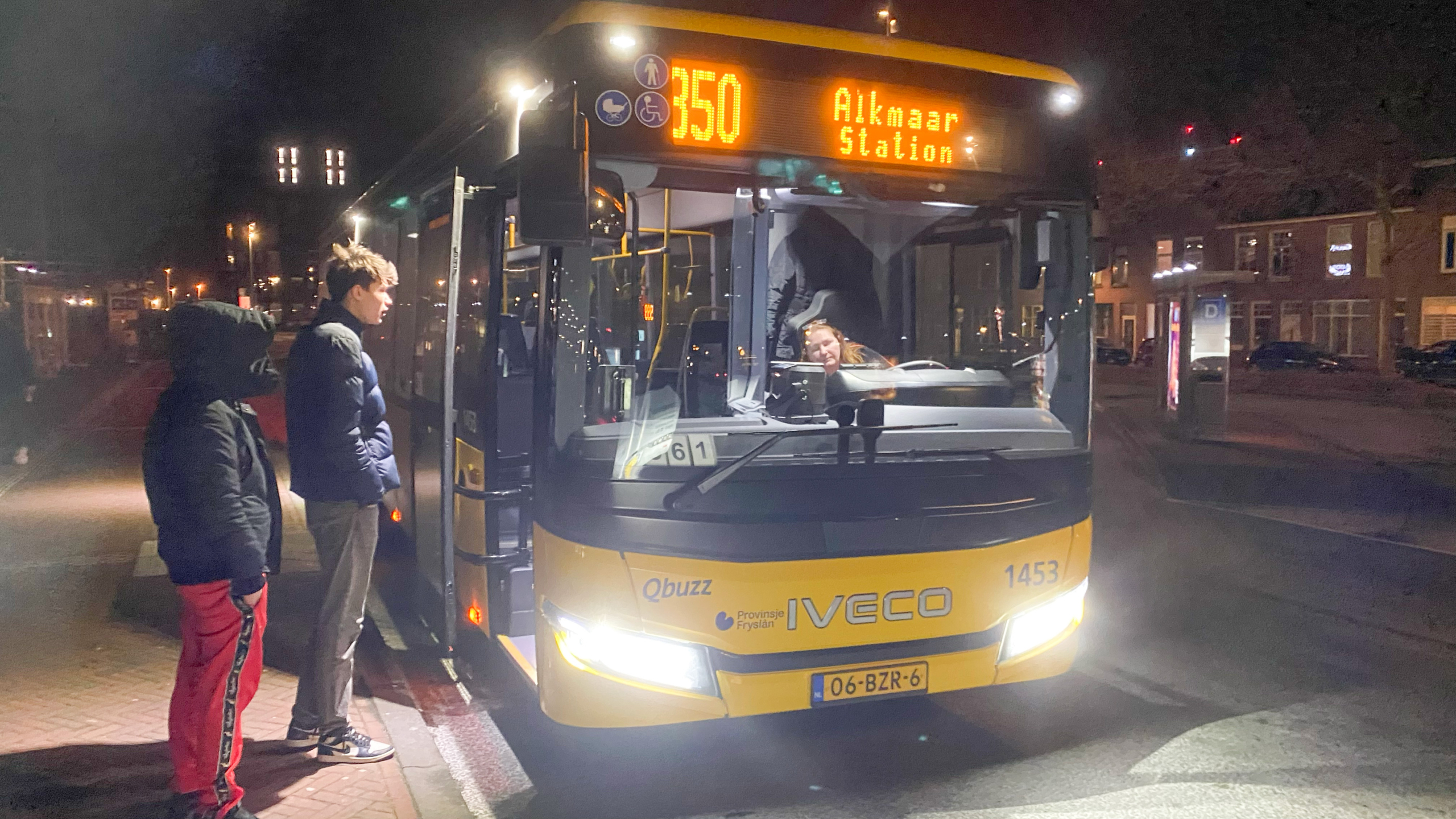 Twee mensen staan op een donkere avond bij een geparkeerde gele bus met de bestemming Alkmaar Station, met het nummer 350 op de voorruit. De bus is van het merk Iveco en heeft het logo van Qbuzz en Provincie Fryslân.