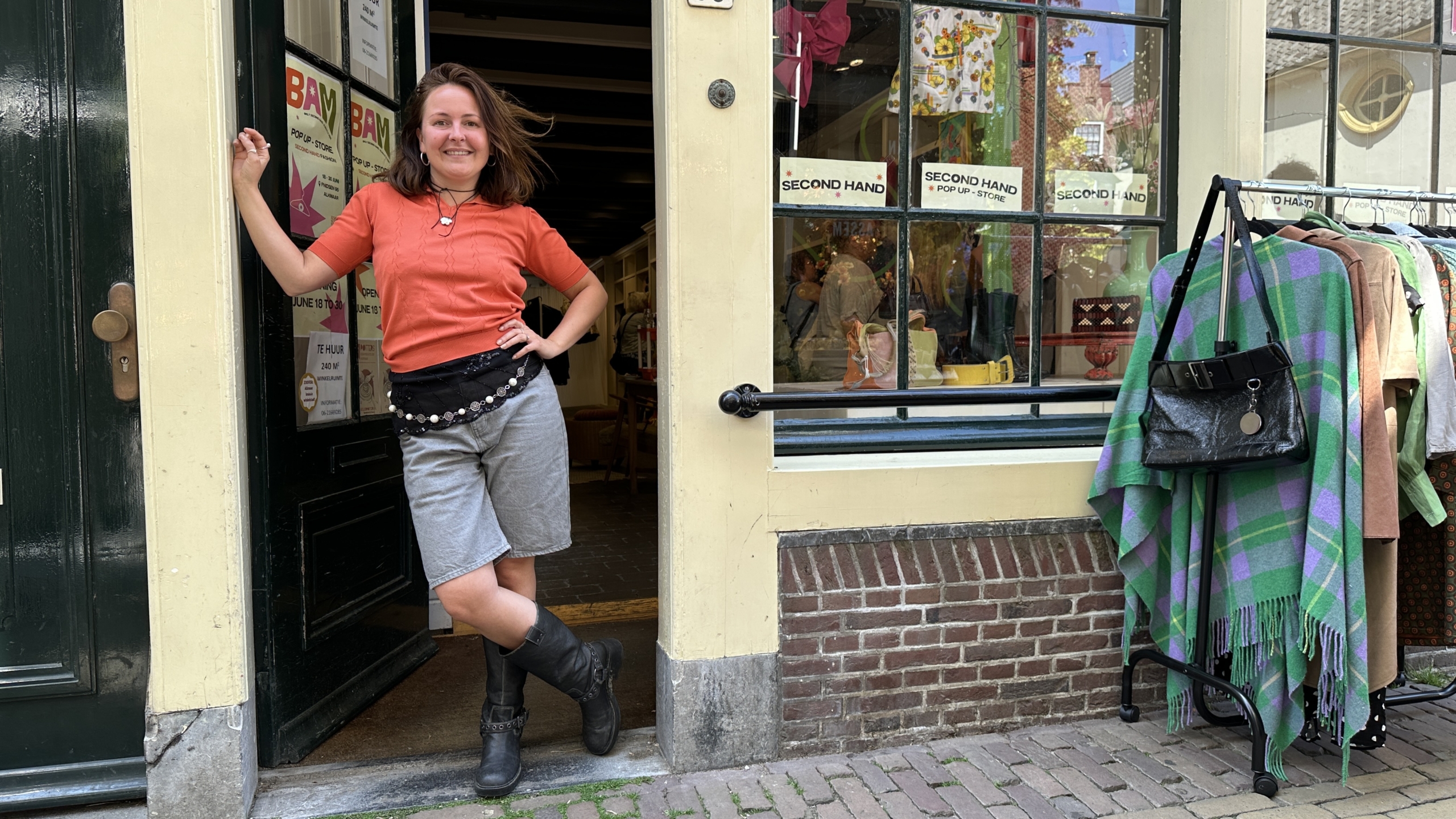Een vrouw in een rode trui en grijze shorts staat in de deuropening van een tweedehandswinkel. Naast haar hangt kleding aan een rek.