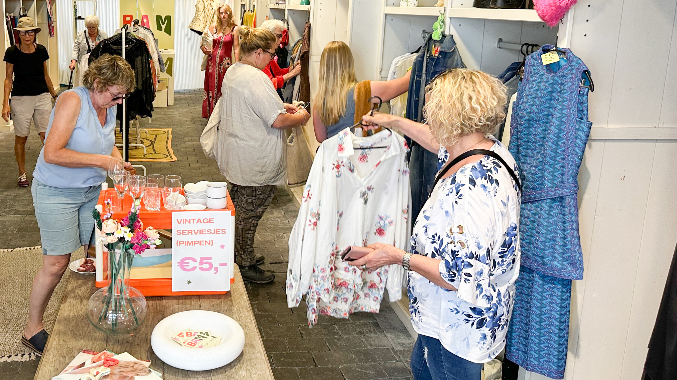 Een vrouw met blond krullend haar houdt een shirt vast in een tweedehandswinkel, omringd door andere klanten die kleding bekijken en snuffelen tussen schappen. Op een tafel vooraan staat een bordje met "Vintage Serviesjes Pimpen" en een prijs van €5.