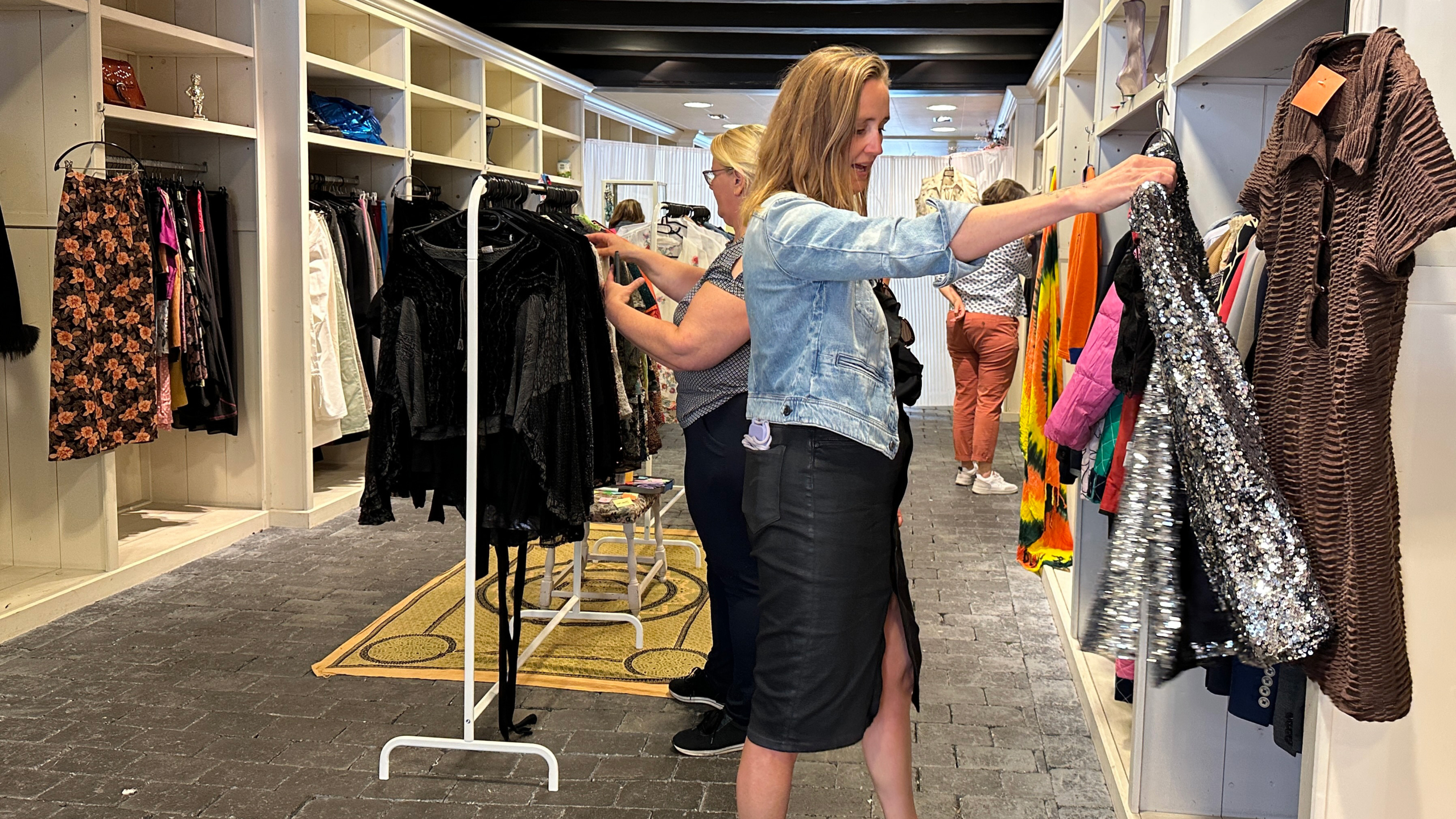 Twee vrouwen winkelen in een kledingwinkel, waarbij de voorste vrouw een sprankelende zilveren jurk bekijkt, omgeven door rekken met diverse gekleurde kledingstukken in een lichte en ruime boetiek.