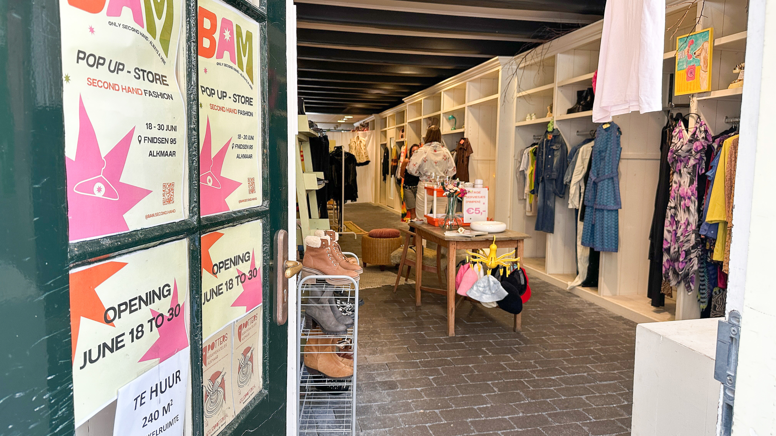 Een open deur naar een pop-up winkel genaamd BAM, met tweedehands mode in Alkmaar, met diverse kledingstukken en schoenen te koop binnenin.