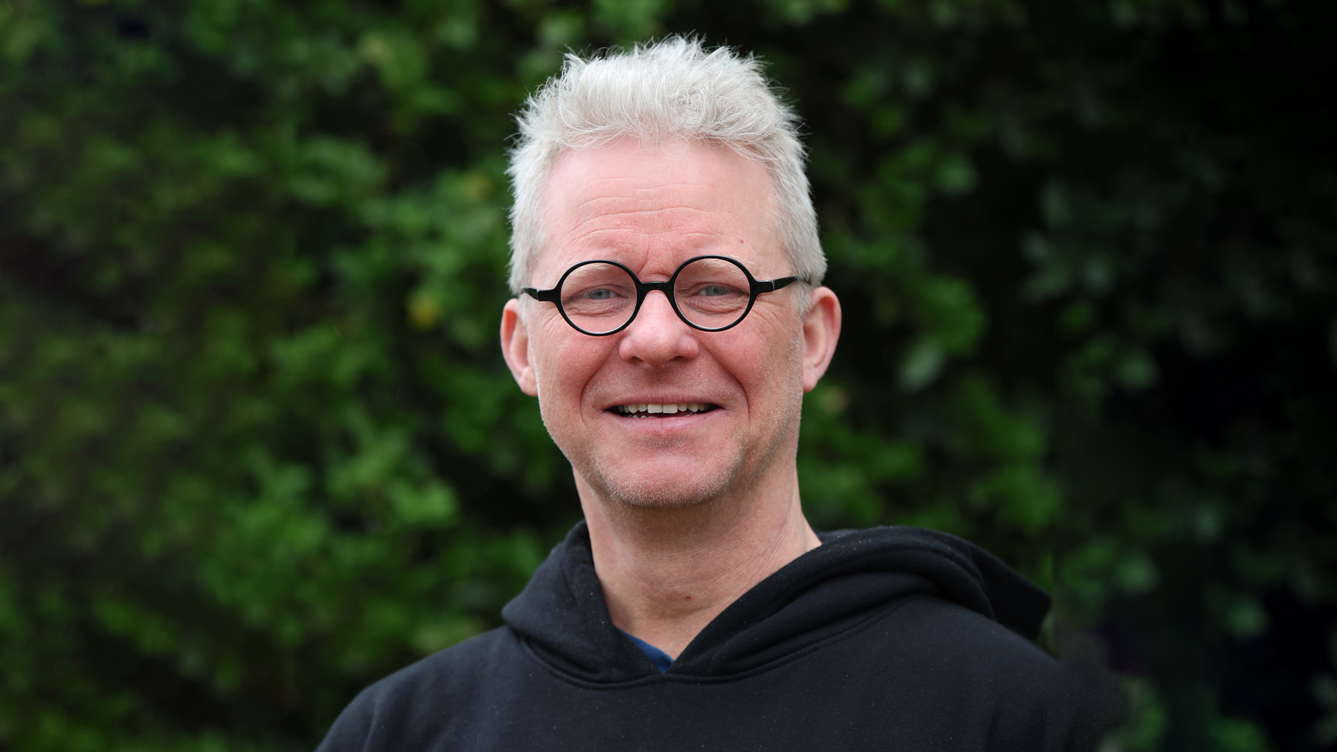 Man met bril en grijs haar draagt een zwarte trui, met een groene achtergrond van gebladerte.