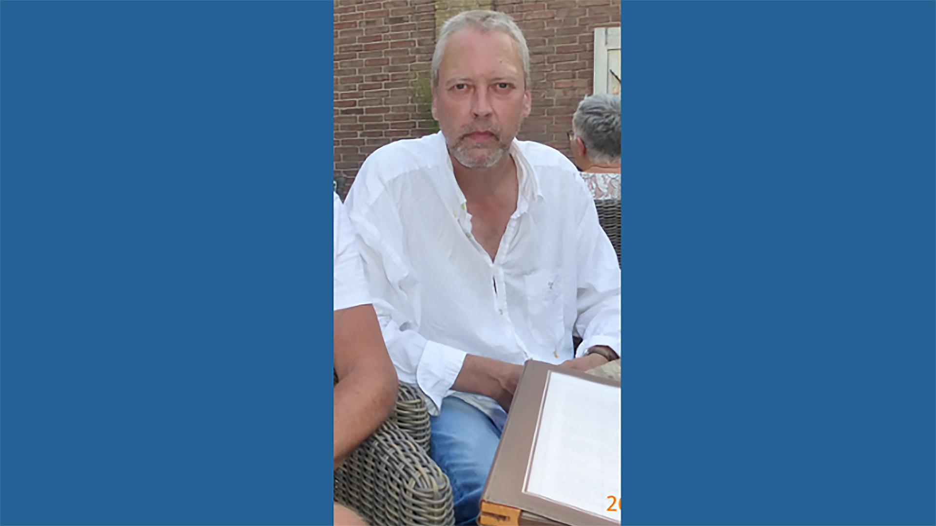 Man in a witte overhemd zit buiten aan een tafel met een menu, tegen een achtergrond van een bakstenen muur.