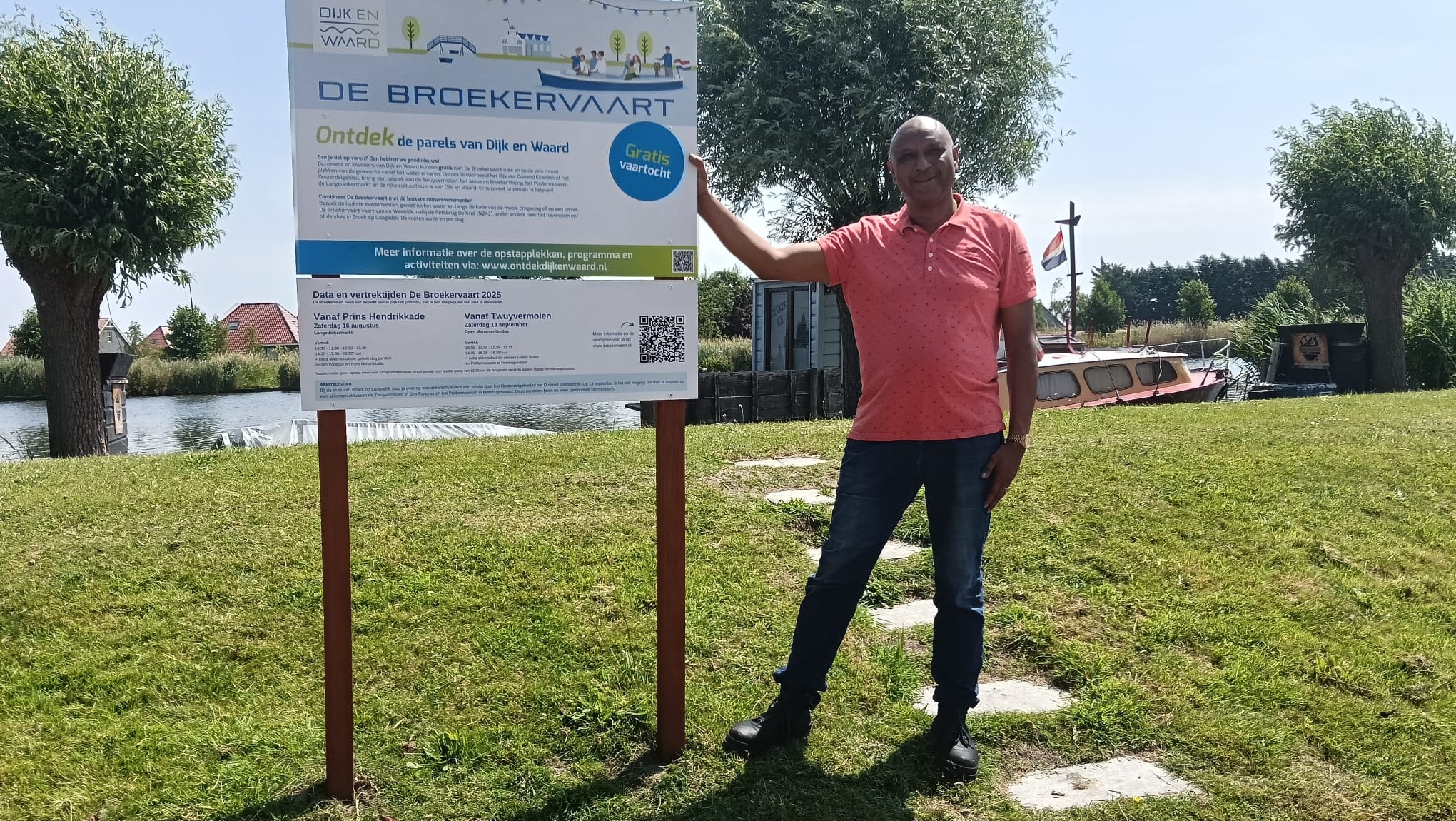 Een man in een roze overhemd staat naast een informatiebord voor 'De Broekervaart' met op de achtergrond een rivier, bomen, en een boot met een Nederlandse vlag.