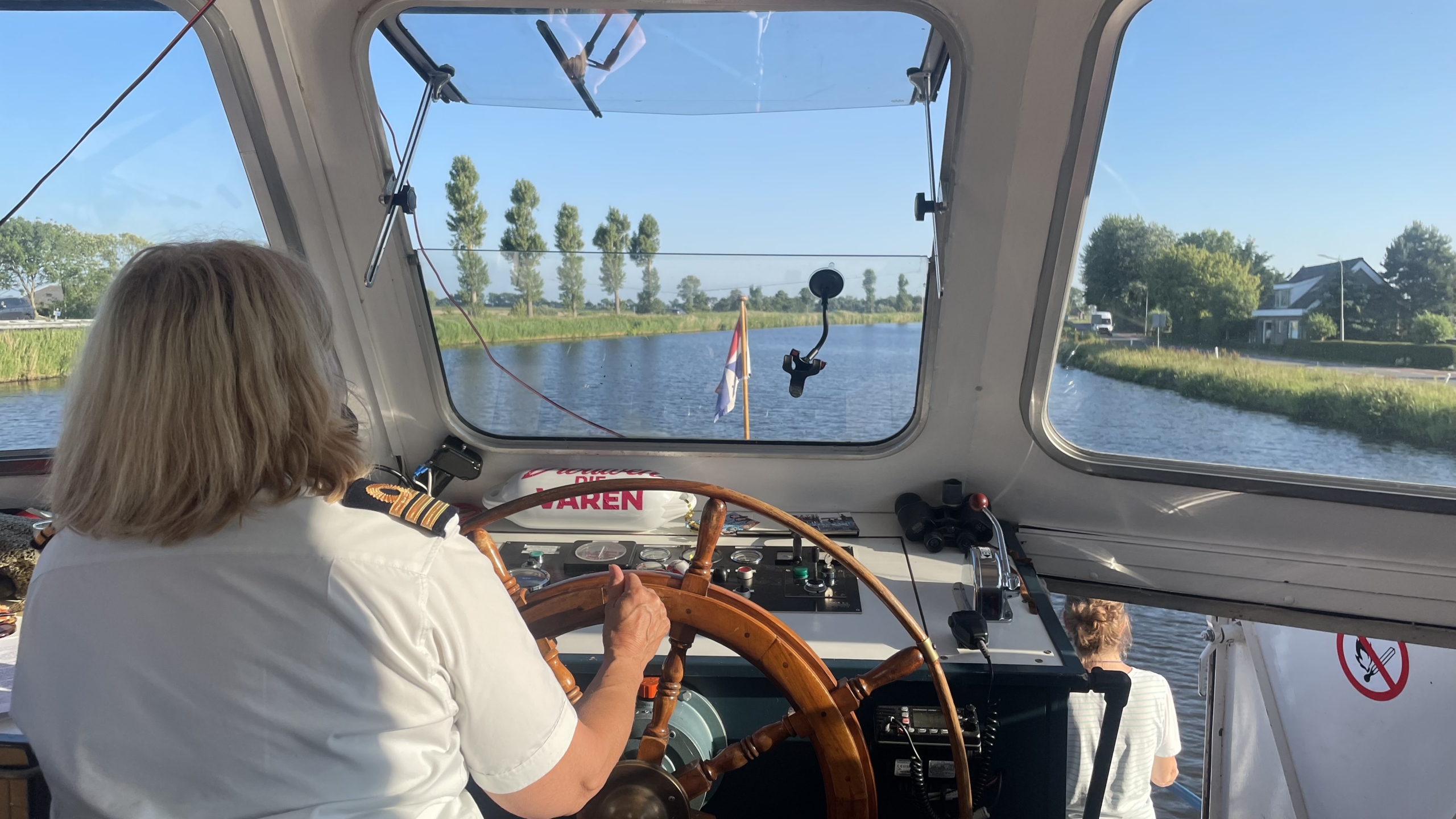 Een vrouw in uniform bestuurt een boot op een kanaal, gezien vanuit de stuurhut met uitzicht op het water en de oever met bomen en huizen.