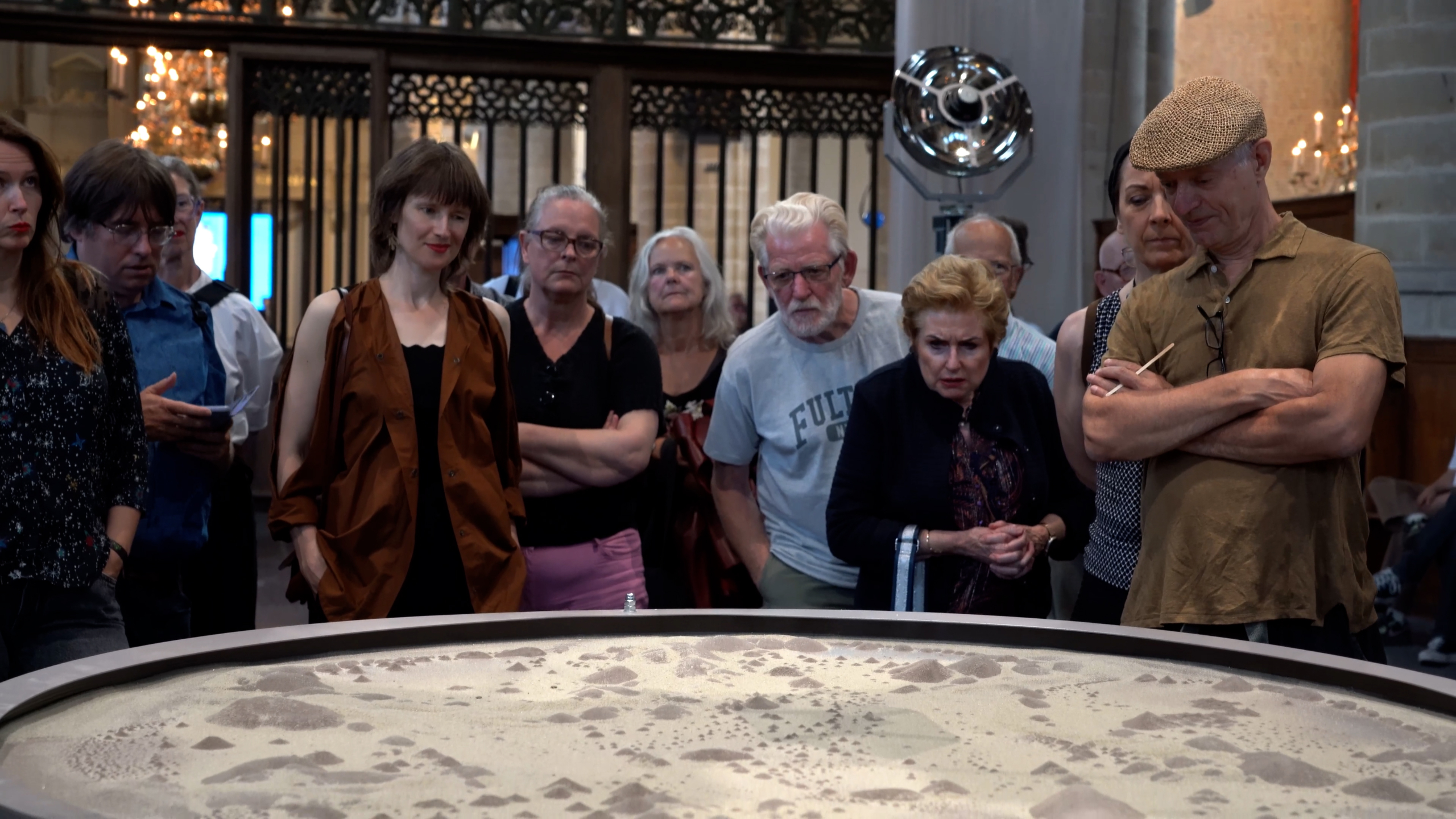 Een groep gevarieerde mensen kijkt aandachtig naar een kunstwerk of installatie in een hal met sierlijke ijzeren hekwerken en een kroonluchter op de achtergrond.