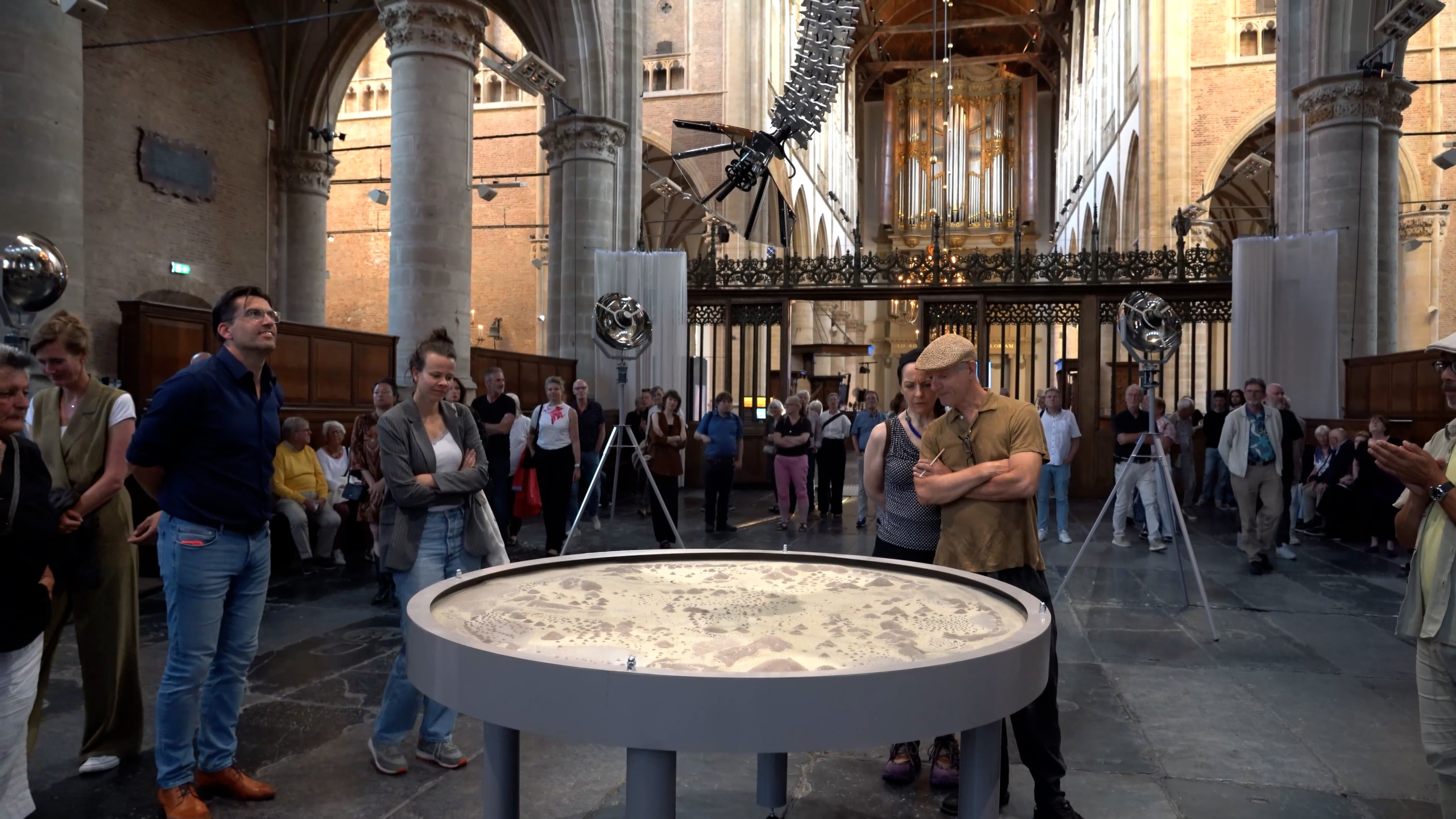 Mensen verzamelen zich rond een tentoonstelling van een ronde tafel met een patroon, binnen in een historische kerk met hoge plafonds en pilaren.