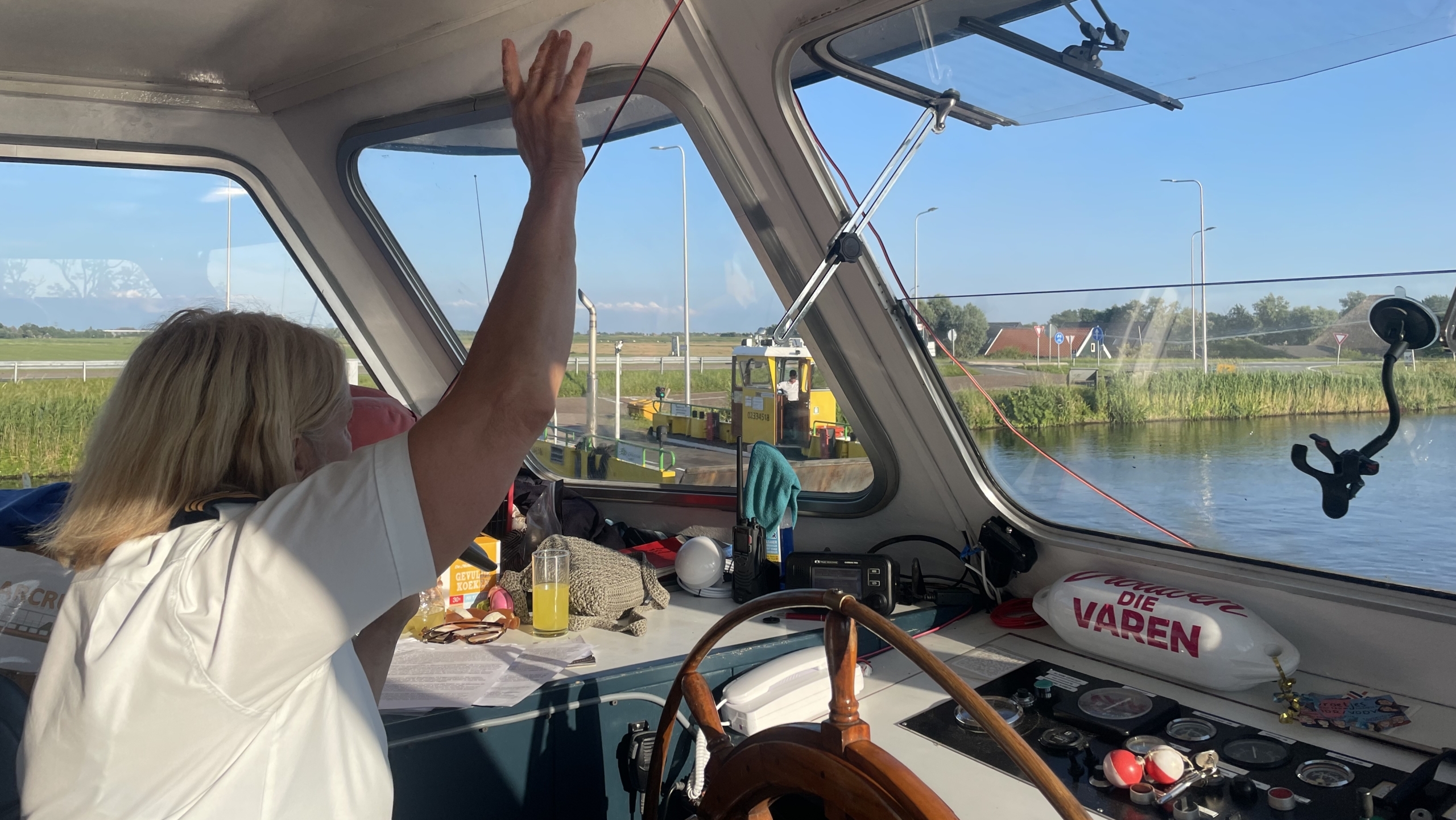 Een persoon in een wit T-shirt bestuurt een boot, met een houten stuurwiel zichtbaar in de cabine. De persoon kijkt naar buiten en heft een hand op. Op de achtergrond is een waterweg zichtbaar met een brug en weelderig groen landschap.