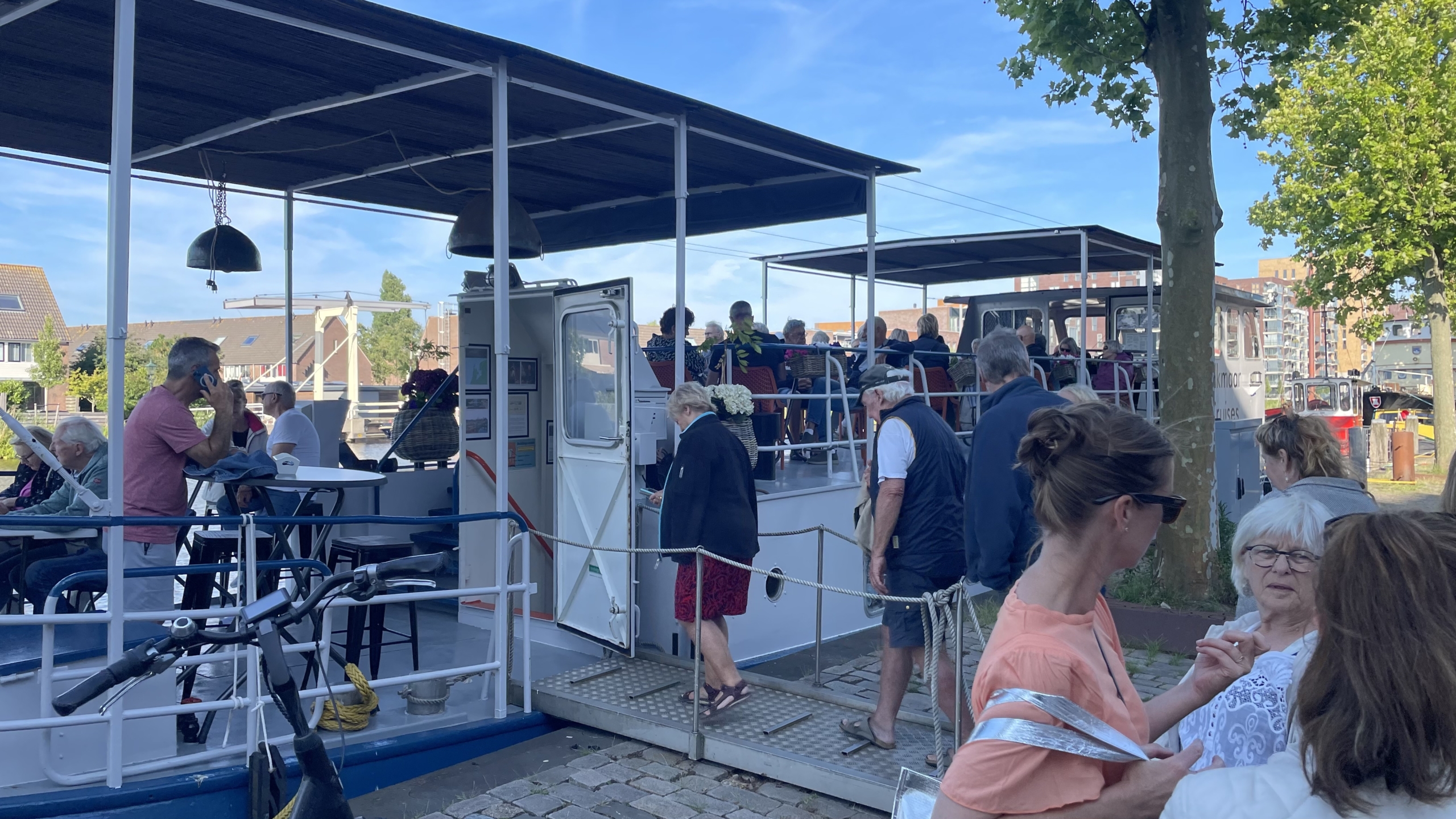 Mensen stappen aan boord van een boot langs een kade, terwijl anderen aan dek zitten. De omgeving toont een stedelijk gebied met bomen en gebouwen op de achtergrond onder een blauwe lucht.
