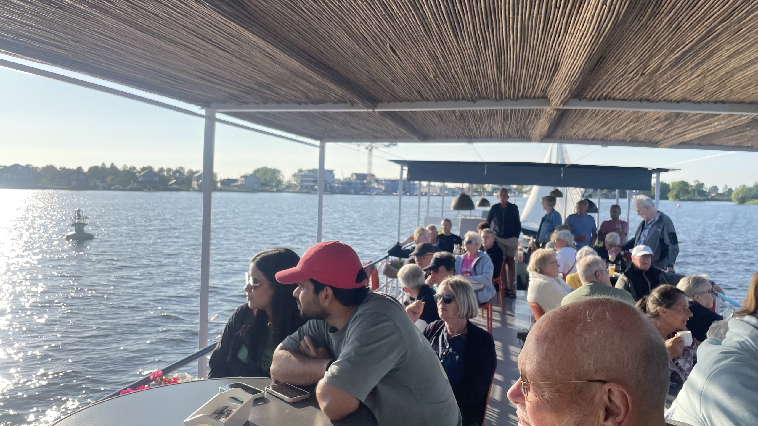 Een groep mensen zit en staat op een overdekte boot op het water op een zonnige dag.
