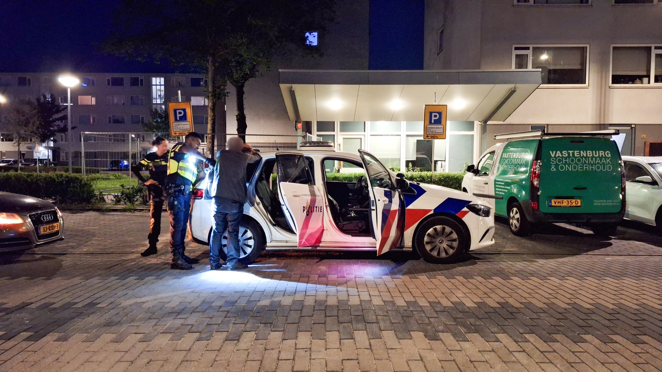 Twee politieagenten controleren een persoon naast een geparkeerde politieauto op een verlichte straat in de avond.