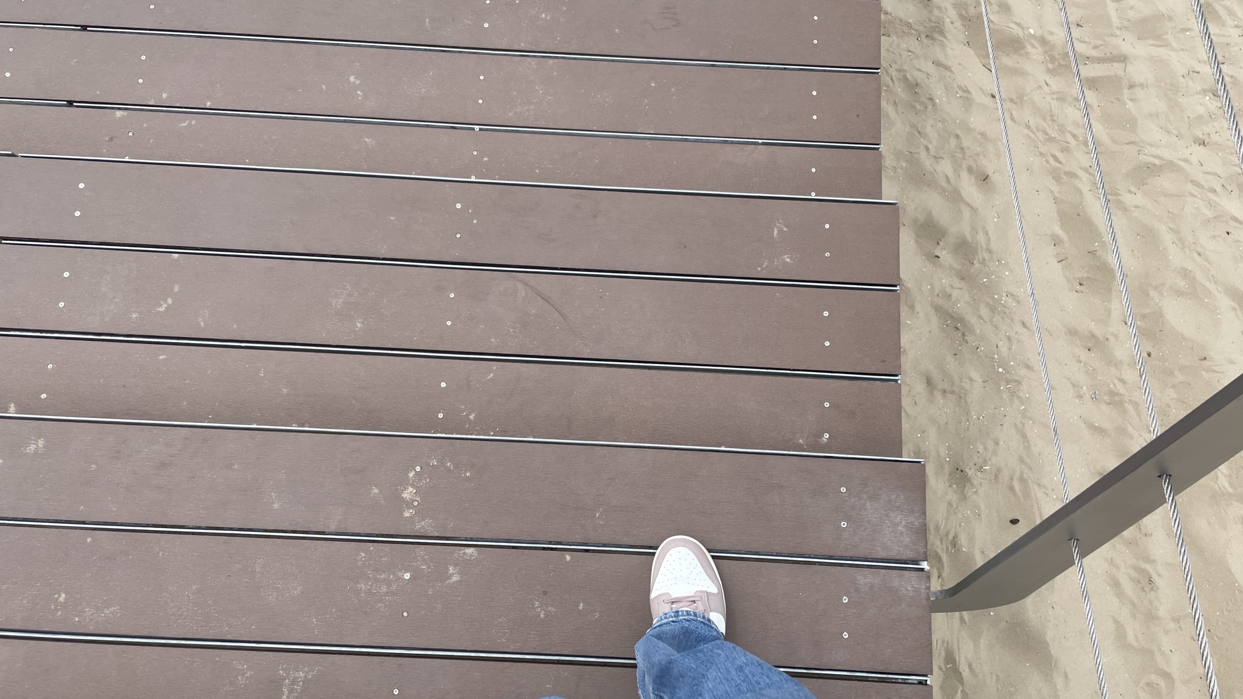 Een persoon met blauwe jeans en witte sneakers staat op een houten vlonder naast een zandgebied met metalen kabels als reling.