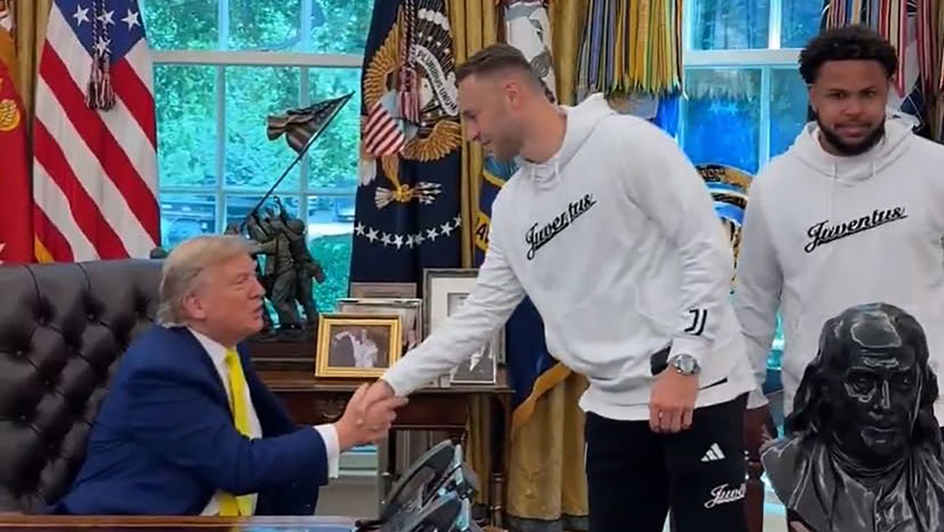 Een man in een blauw pak en gele stropdas schudt de hand van een man in een witte Juventus trui, terwijl een andere man in een vergelijkbare trui erbij staat in een kantoor met Amerikaanse vlaggen en een borstbeeld.