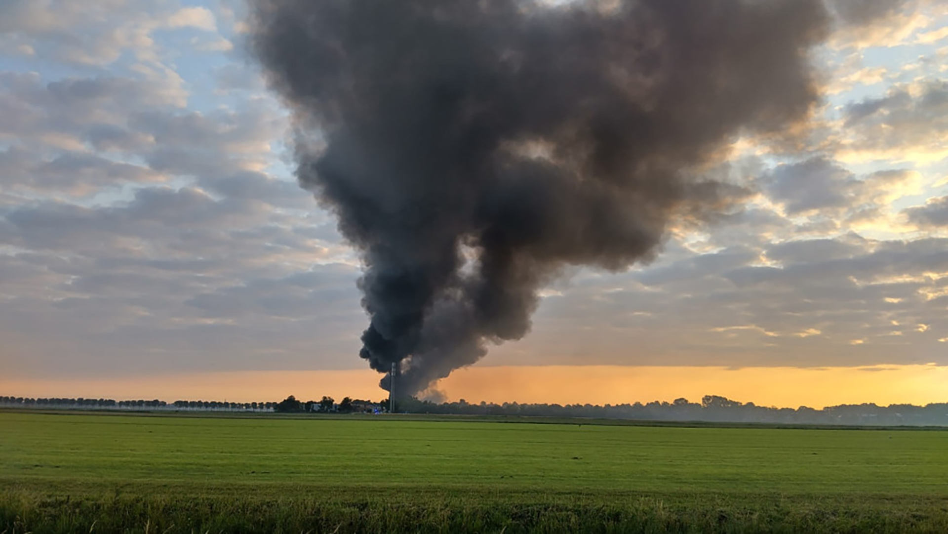 Grote zwarte rookwolken stijgen op vanaf een brand in de verte in een open veld bij zonsondergang, met een gedeeltelijk bewolkte hemel.